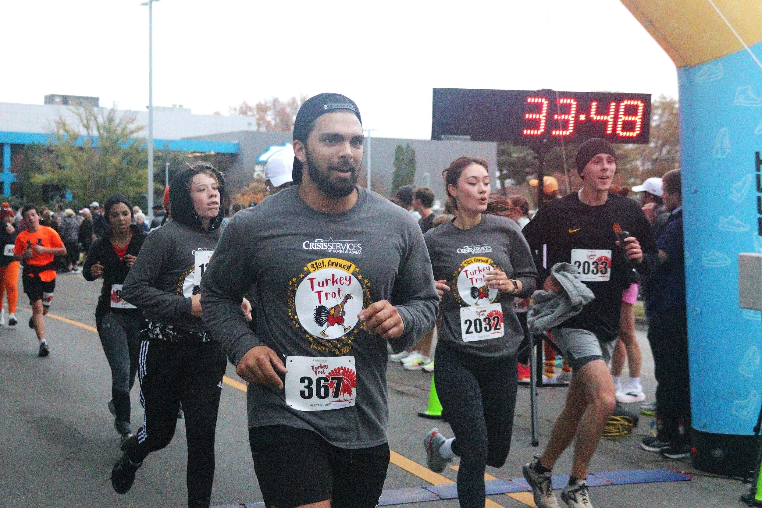 Runners participating in the 31st annual Turkey Trot race, nearing the finish line at 33 minutes and 48 seconds, outdoors on a city street with a large digital clock and a blue finish line arch.