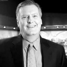 A black and white photo of a man smiling in an indoor setting, wearing a suit and tie.