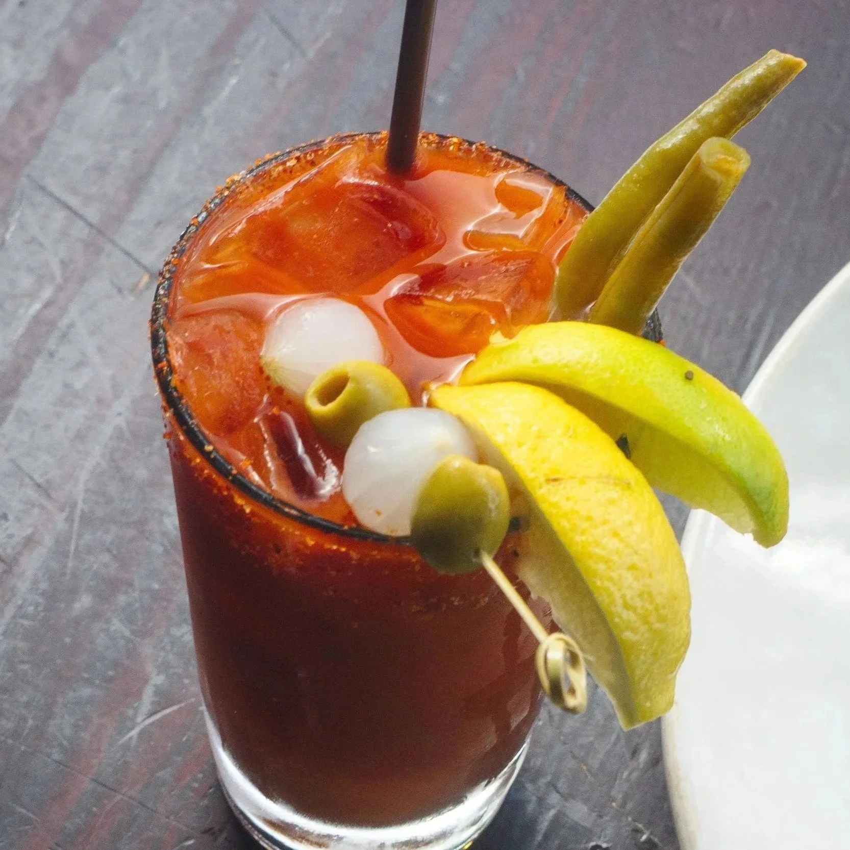 A tall glass of Bloody Mary garnished with lemon wedges, green olives, and pickles, with a straw in the drink.