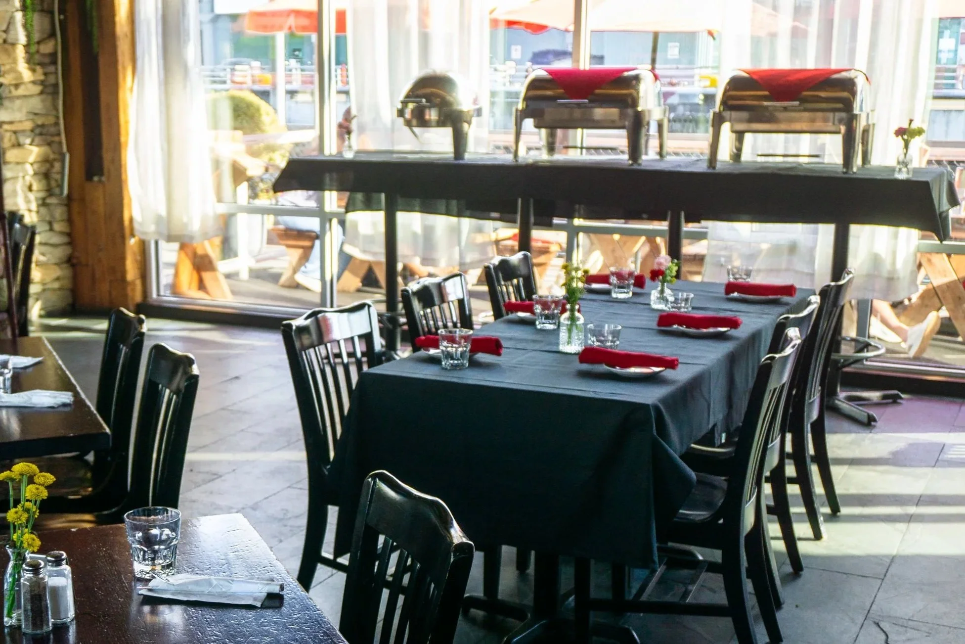 A dining area with a black tablecloth-covered table set with glasses, red napkins, and small floral centerpieces, inside a restaurant with large windows, outdoor seating, and buffet chafing dishes on a table in the background.