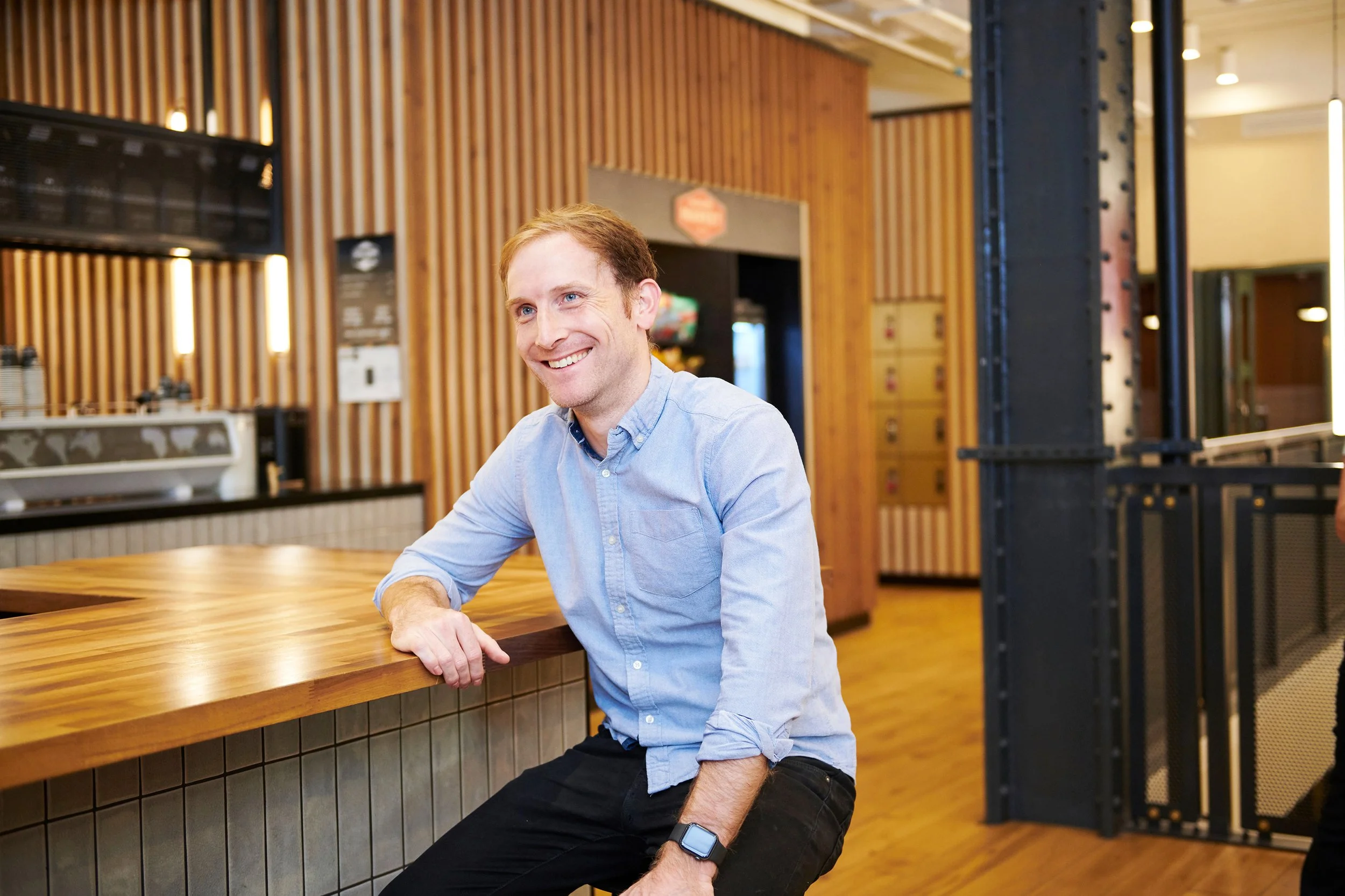 A man with light brown hair and a blue button-up shirt sitting at a wooden bar counter, smiling in a modern, warmly lit cafe or restaurant.