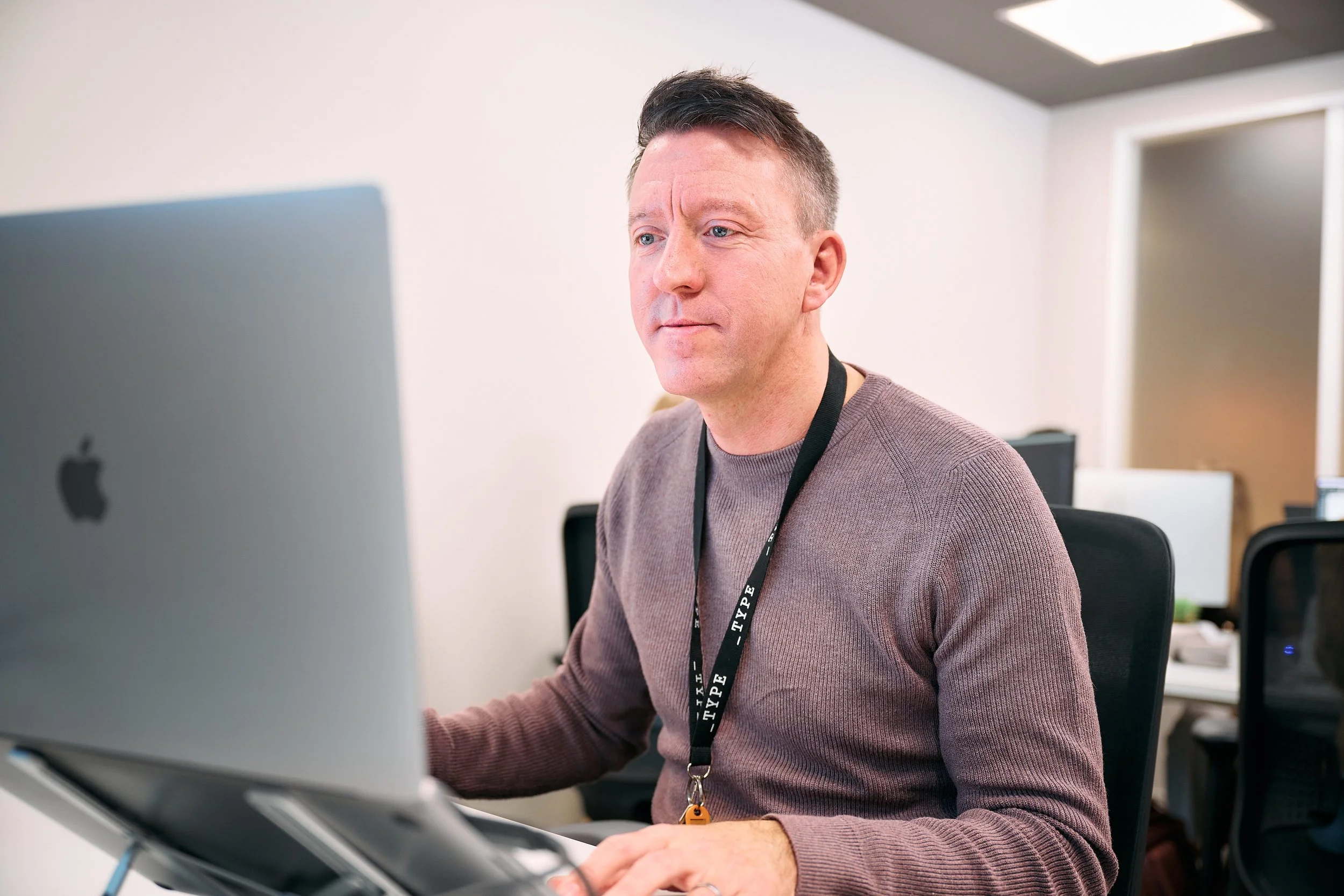 A man with short dark hair, wearing a brown sweater and a black lanyard, sitting at a desk working on a silver Apple iMac computer in an office environment.