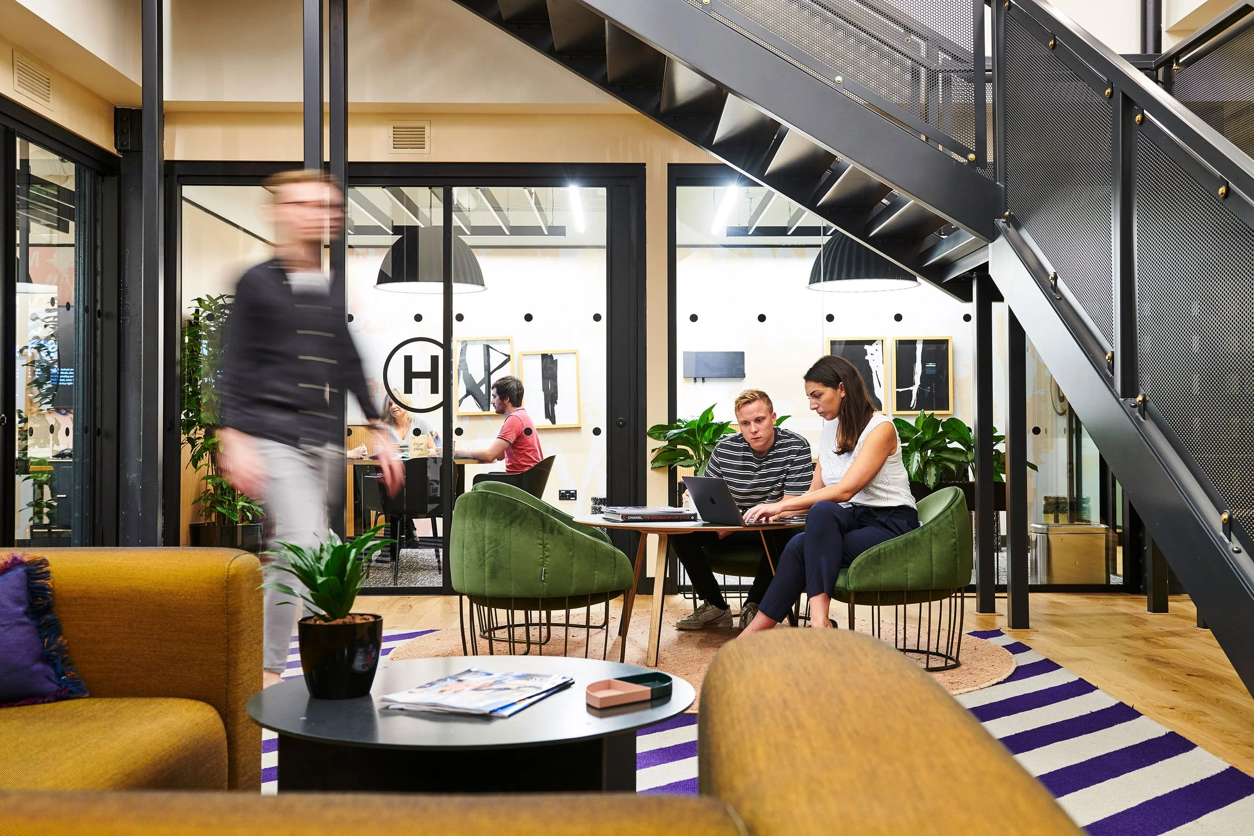 People working and walking inside a modern, open-concept office with glass walls, green chairs, plants, and artwork.