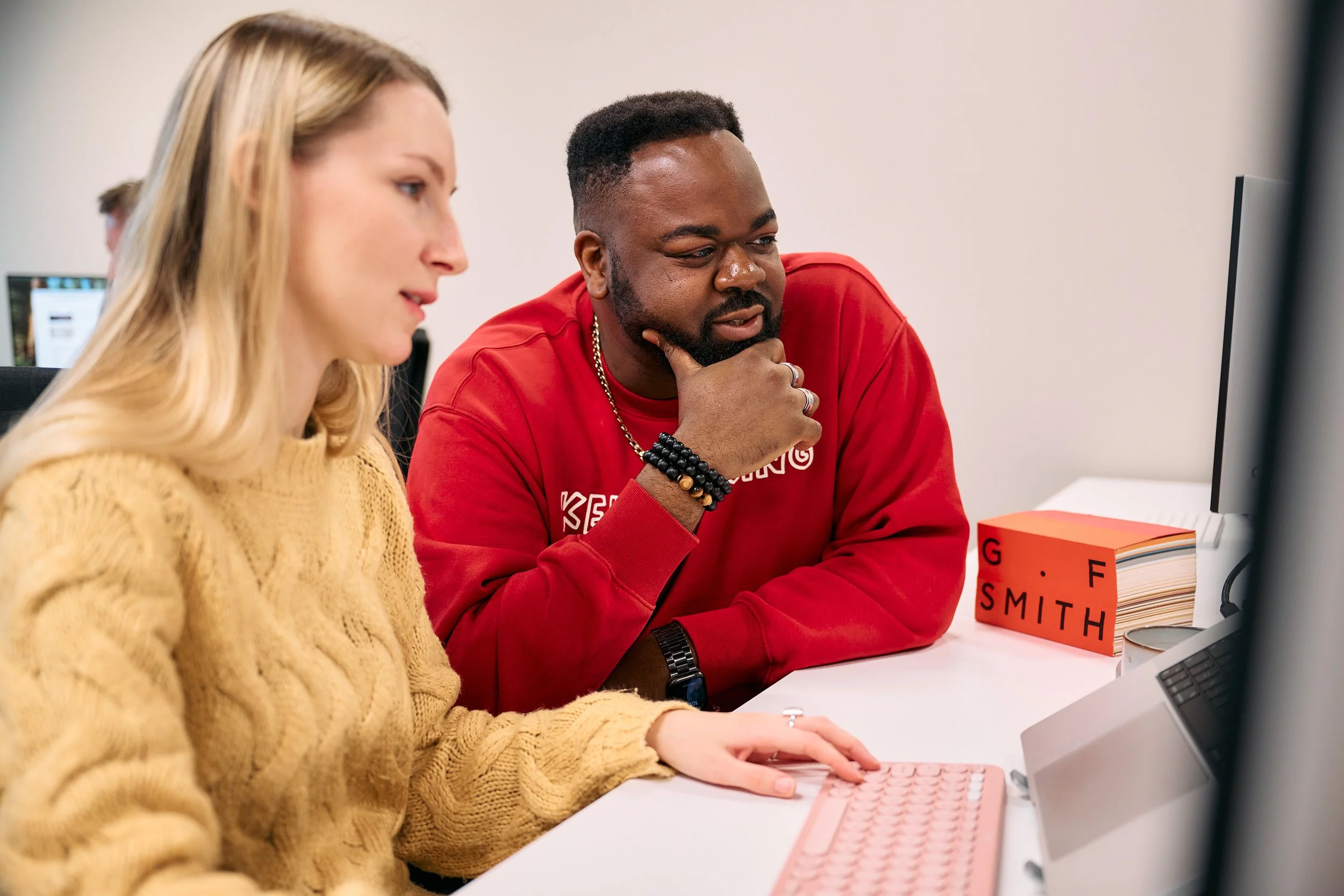 Two people working together at a computer in an office, one woman with blonde hair in a beige sweater and one man with a beard in a red sweatshirt, looking at the monitor.