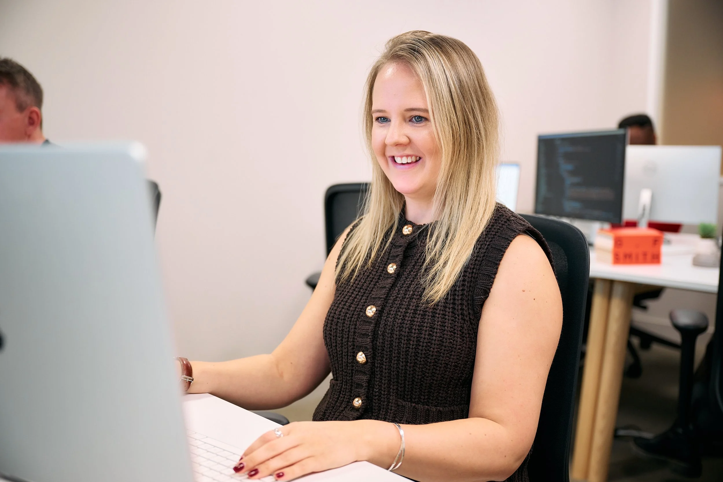 A woman with blonde hair, smiling and working at a computer in an office setting.