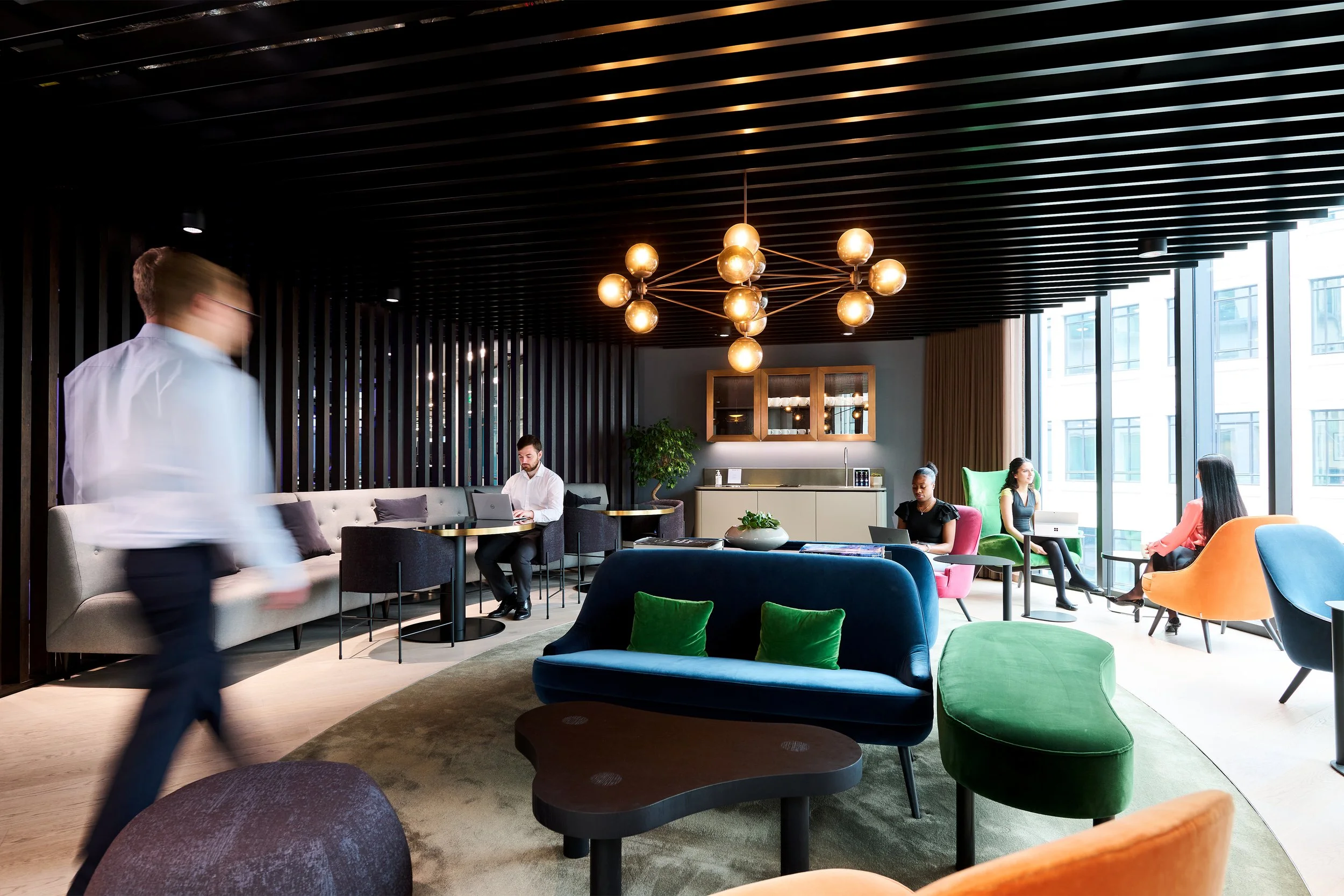 Modern office lounge with colorful chairs, sofas, people working on laptops, and large windows.