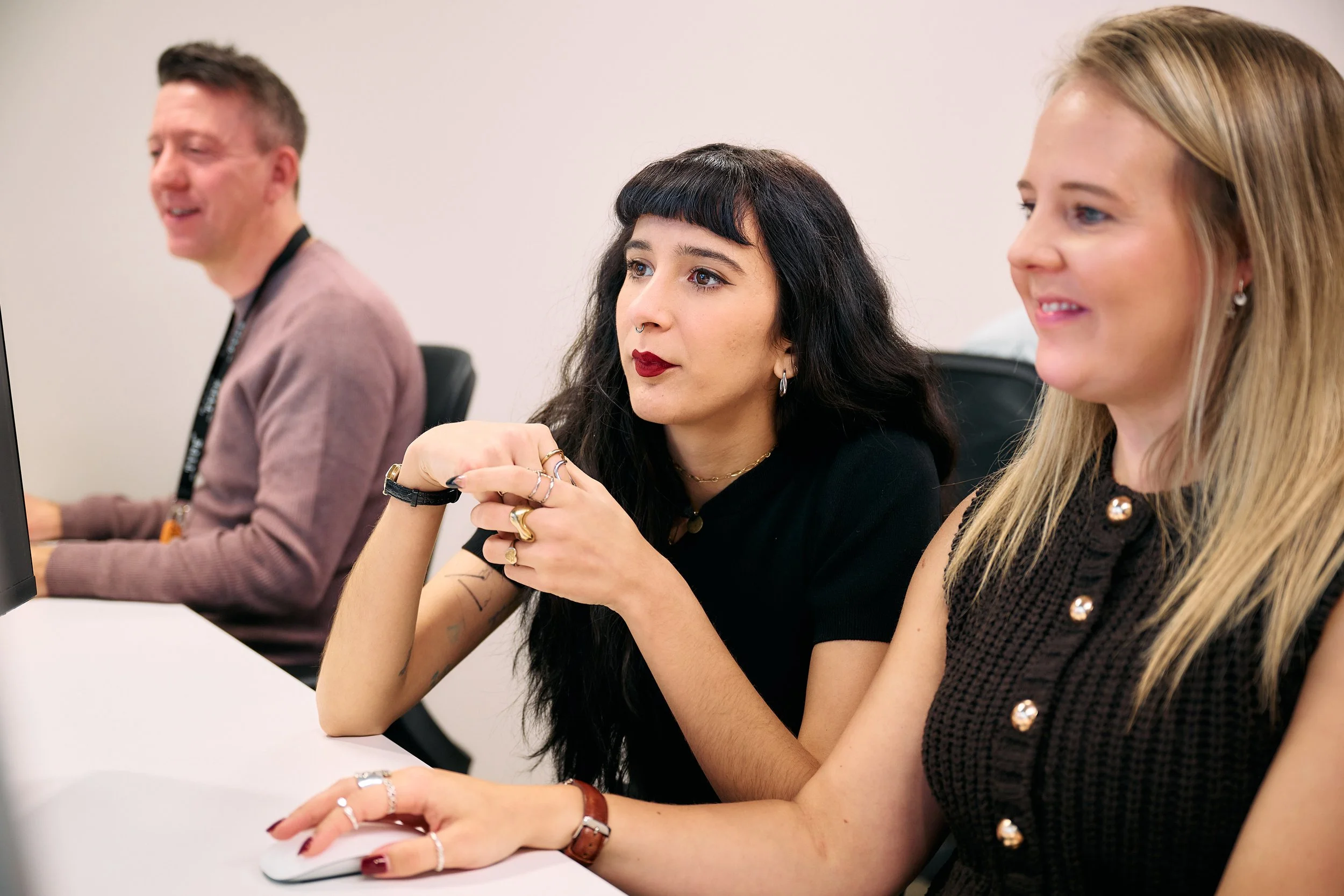 Three people sitting at a table, with the woman on the right working on a computer, the woman in the middle listening, and the man on the left looking forward.