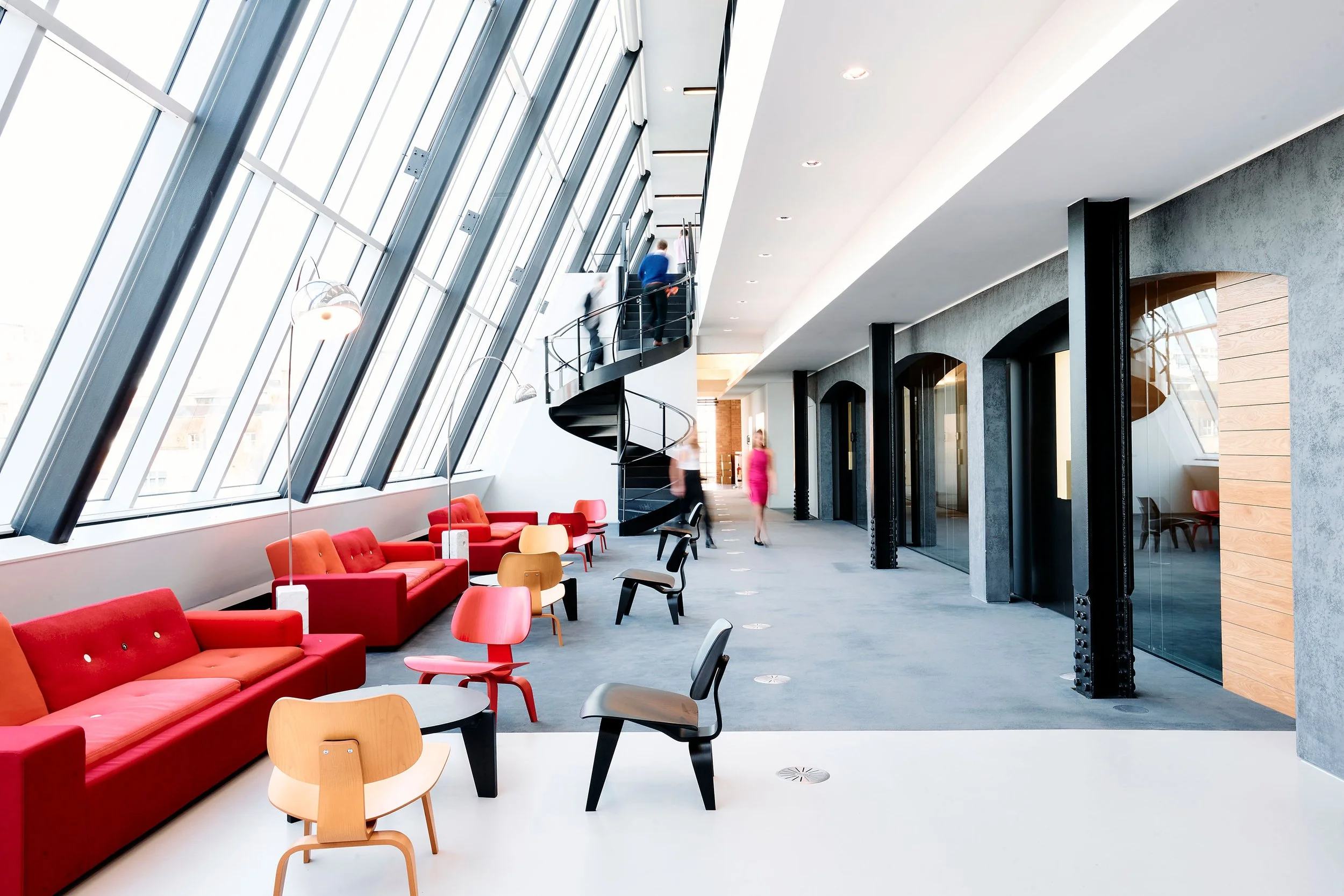 Modern office lobby with large angled windows, red and beige chairs, and a black spiral staircase.