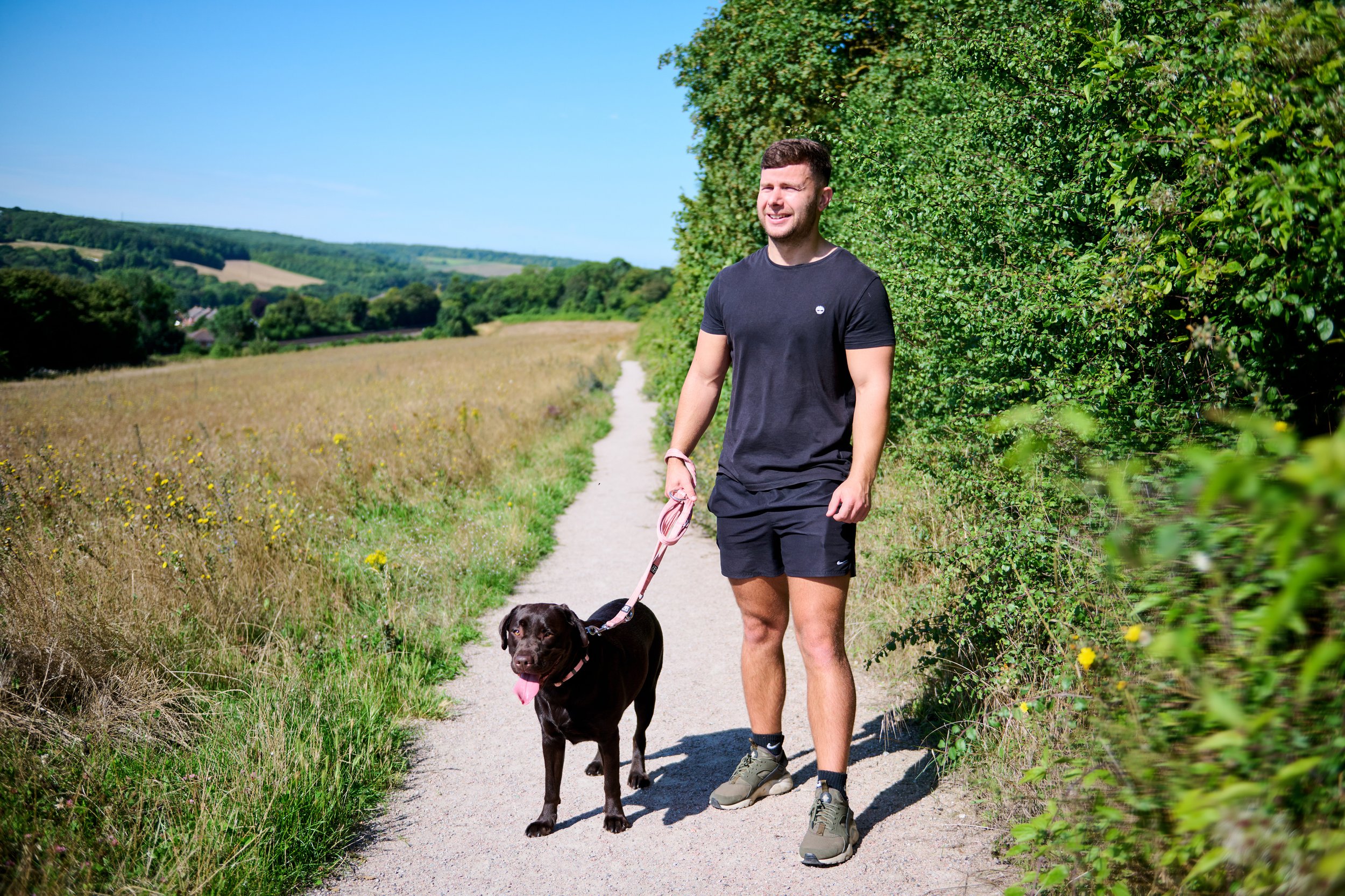 A man walking a black dog on a leash along a rural dirt path in a scenic countryside with green fields and trees on a sunny day.
