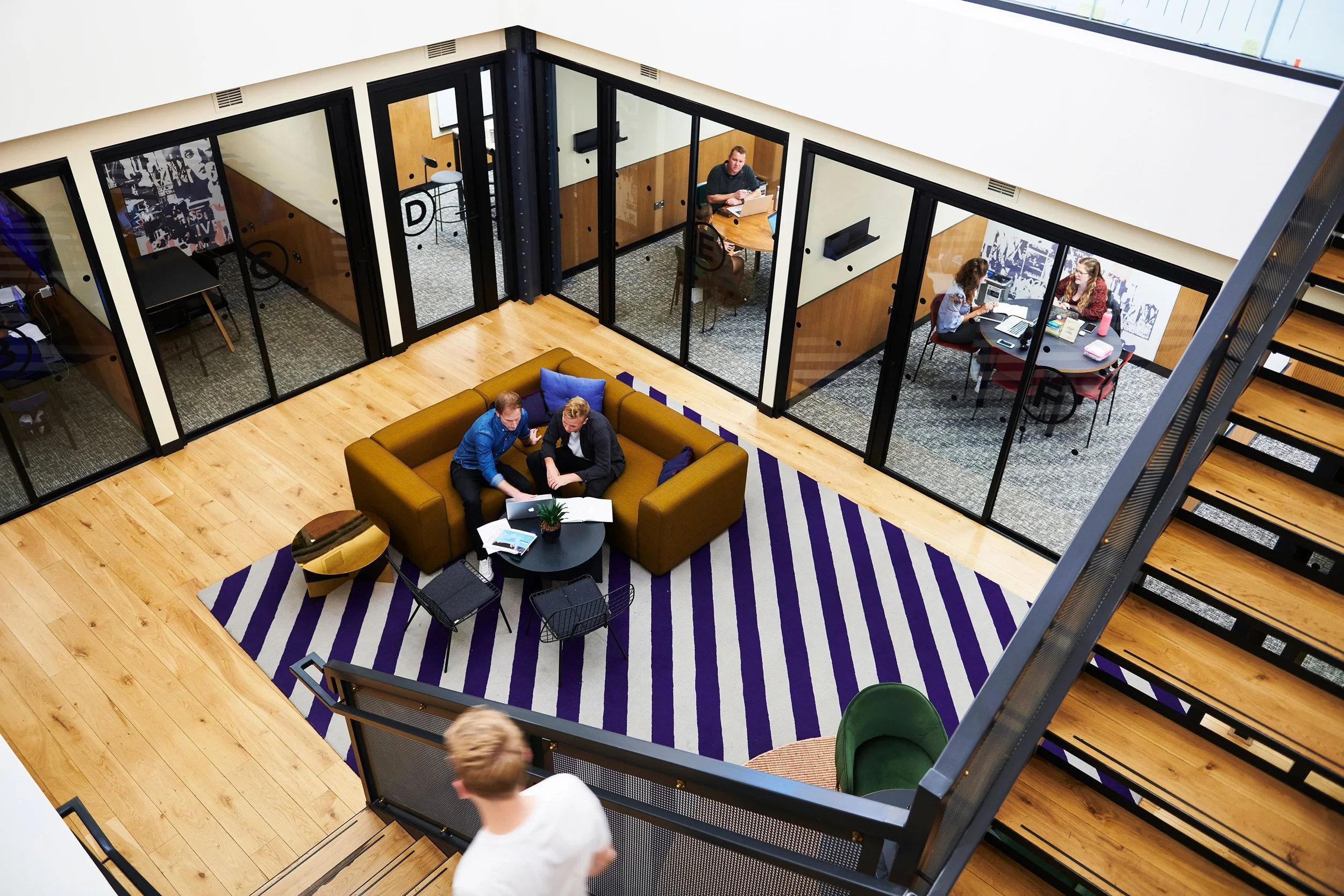 An aerial view of a modern office space showing a central lounge area with two people working on a laptop. Surrounding the lounge are glass-enclosed meeting rooms with people working inside. A staircase is visible on the right side of the image.