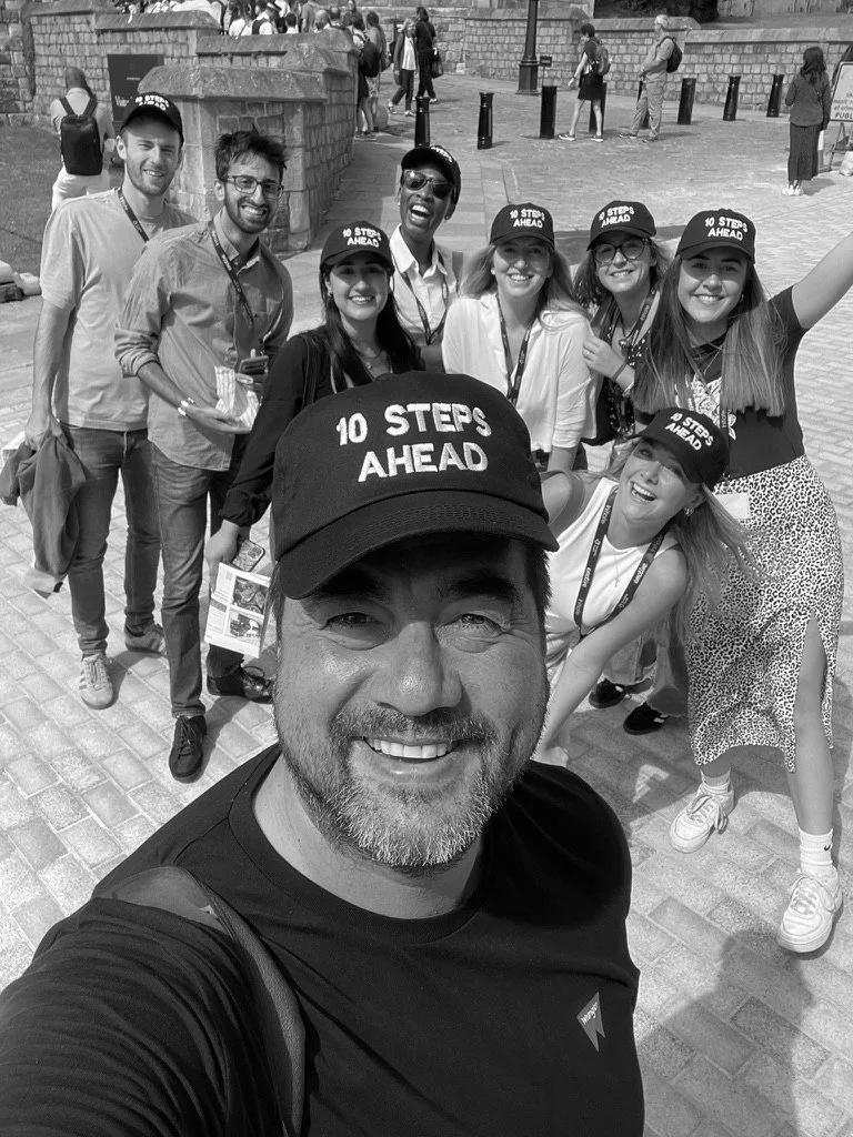 A group of people smiling and posing for a selfie outdoors, with some wearing caps that say "10 Steps Ahead." The man in the foreground taking the selfie is wearing a cap with the same phrase and a black shirt. The group appears to be at a public event or gathering.