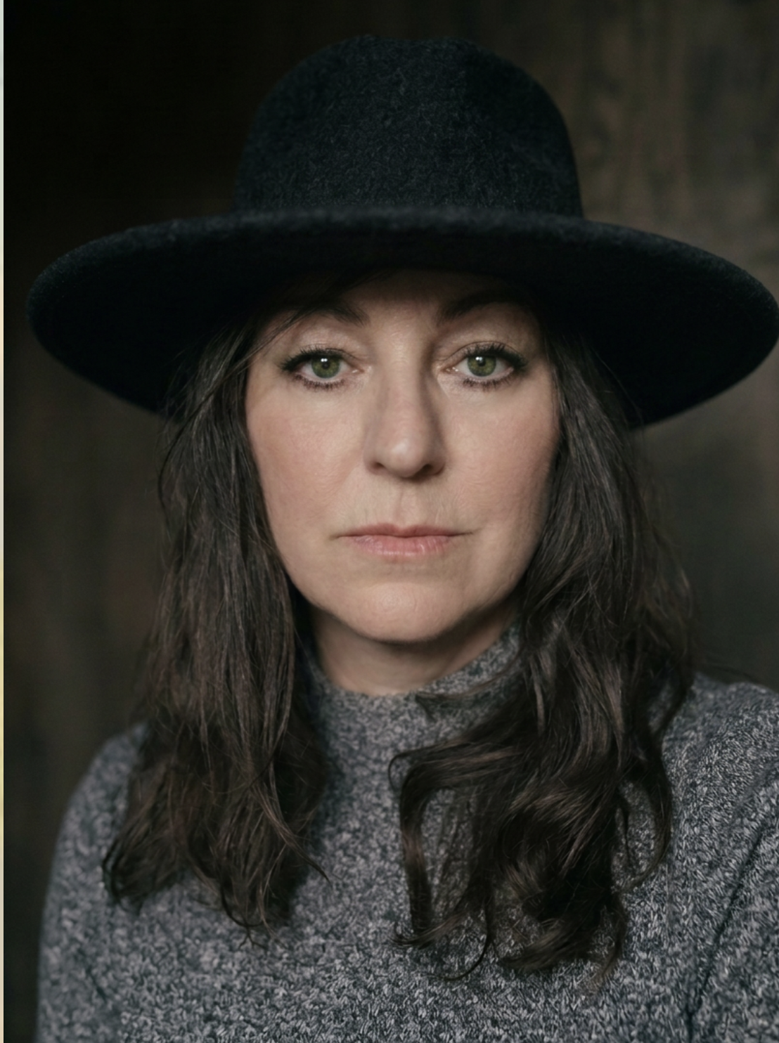 A woman with green eyes and dark wavy hair wearing a black hat and a gray turtleneck sweater.