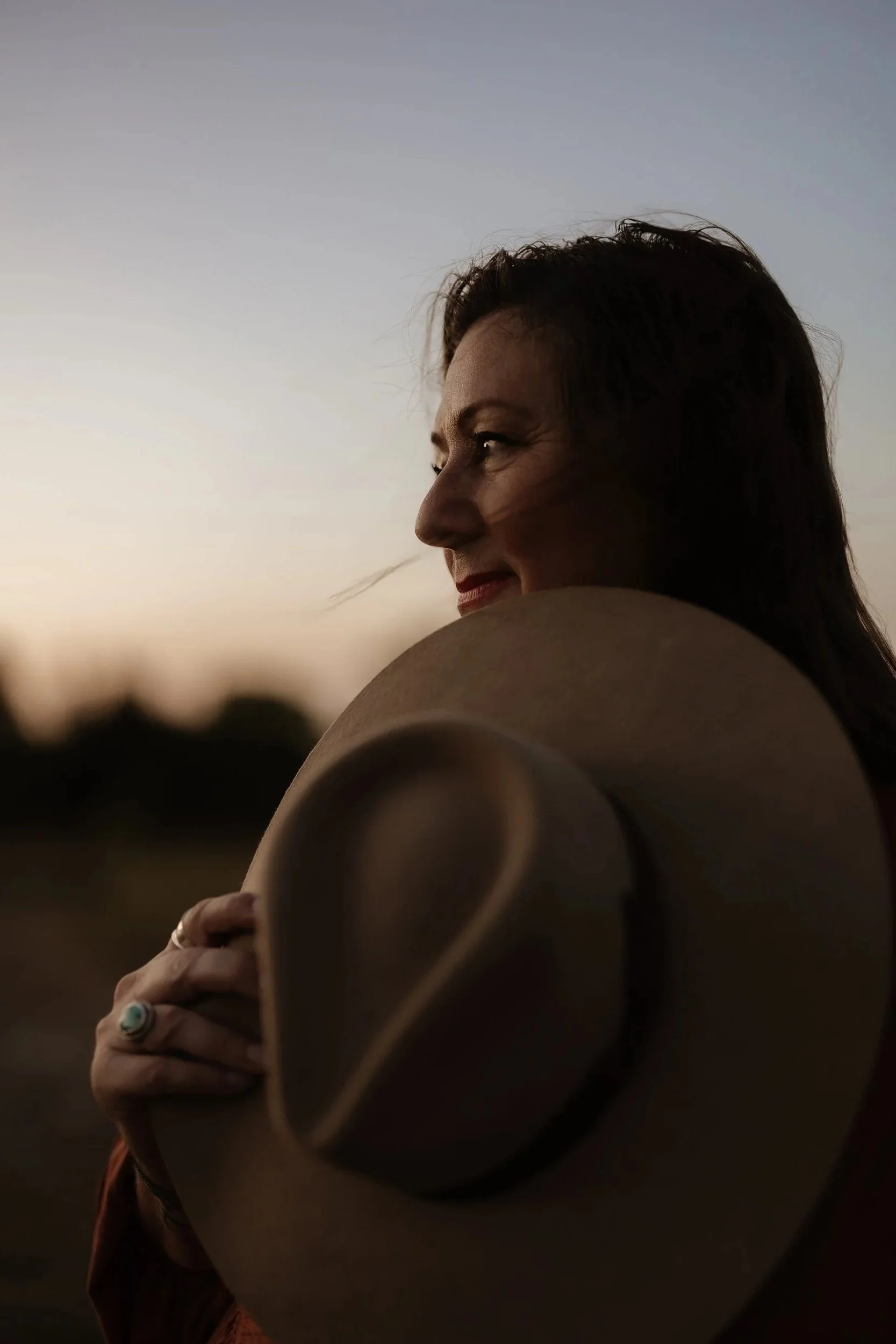 A woman with dark hair holding a western hat near her face during sunset with a blurred outdoor background.