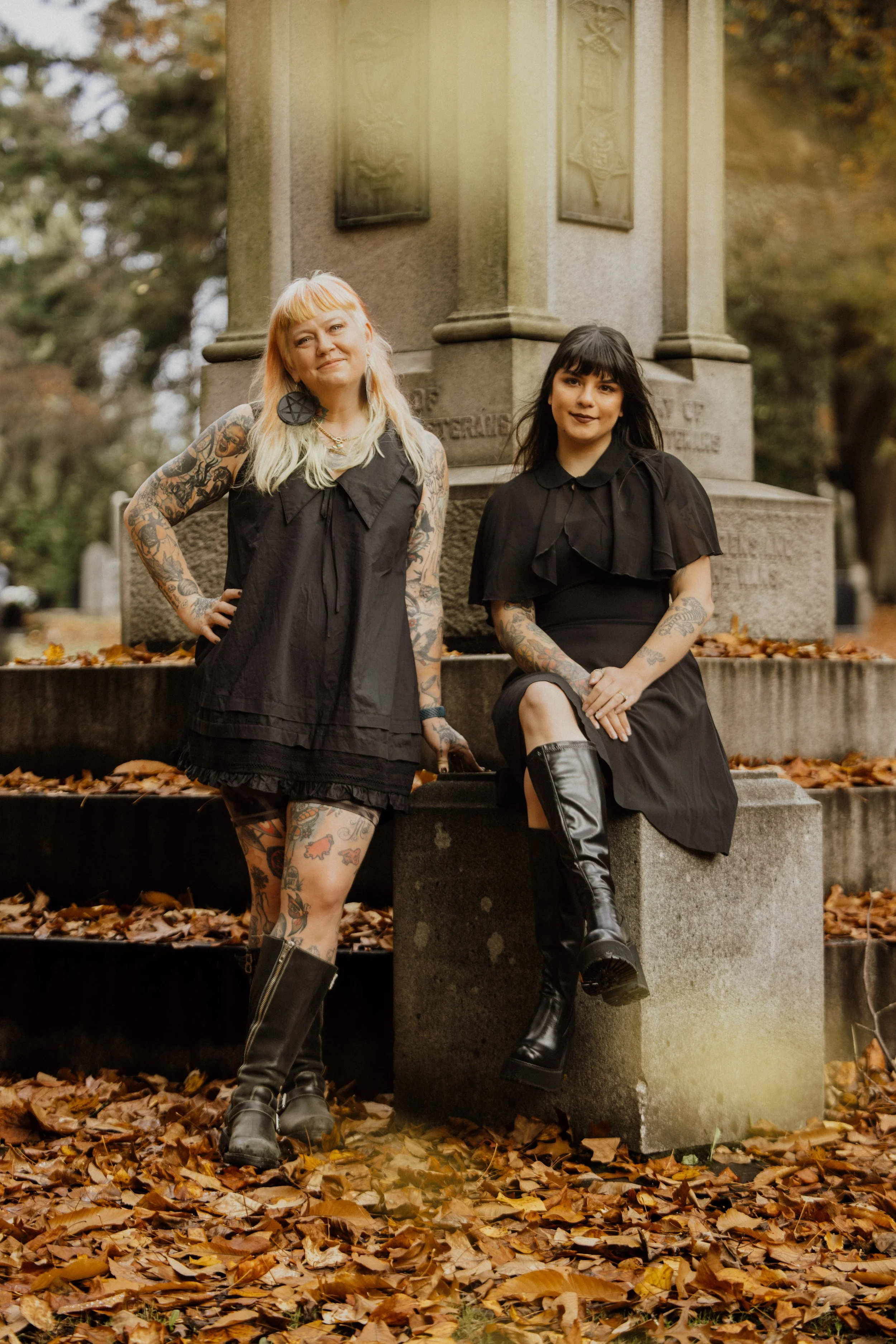 Two women with tattoos, black clothing, and boots pose near a stone monument in a park during autumn.