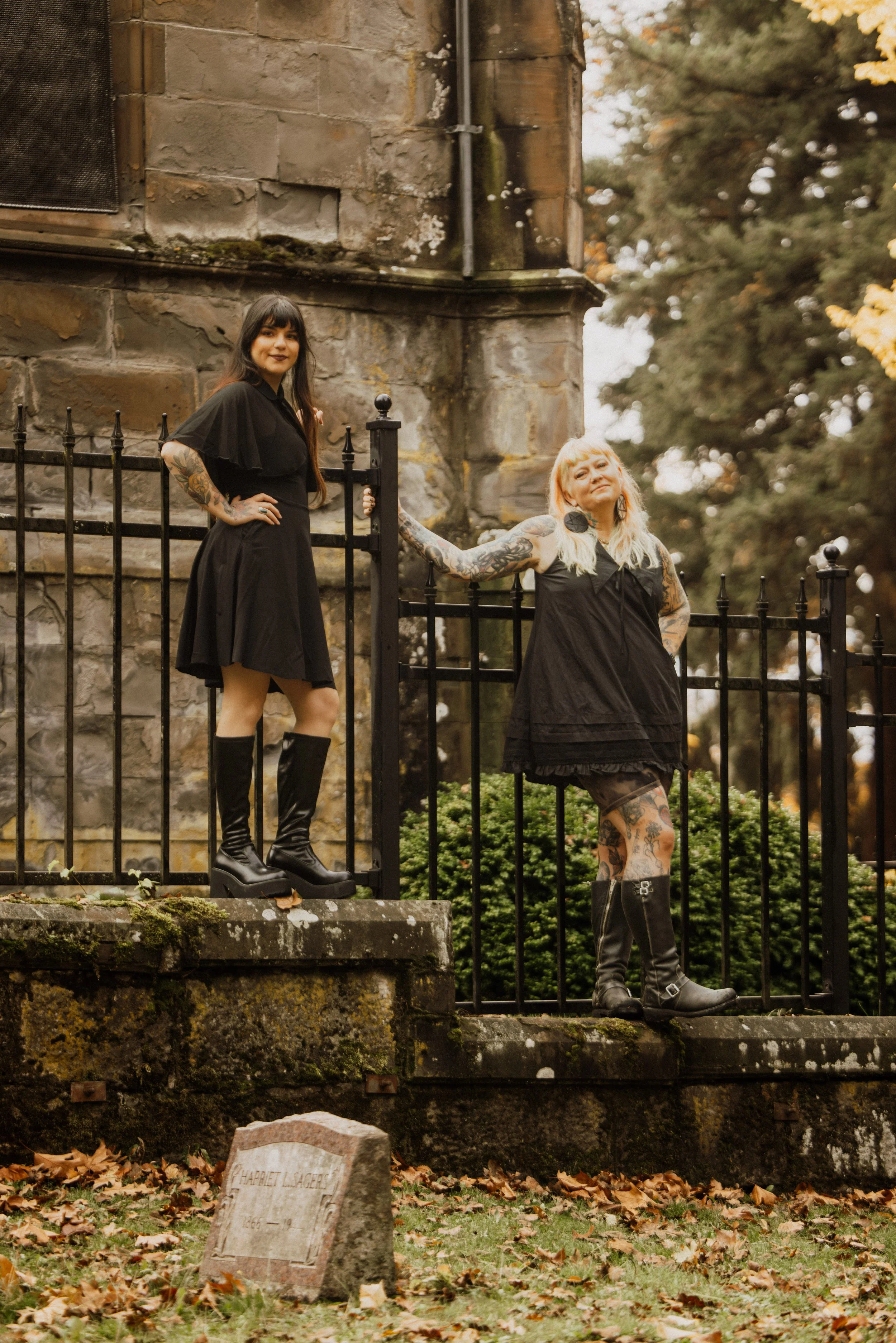 Two women with tattoos standing on a stone ledge by a black wrought iron fence and stone building. One woman is wearing a black dress and tall black boots, and the other is wearing a black dress with tattoos showing and black boots. They are outdoors