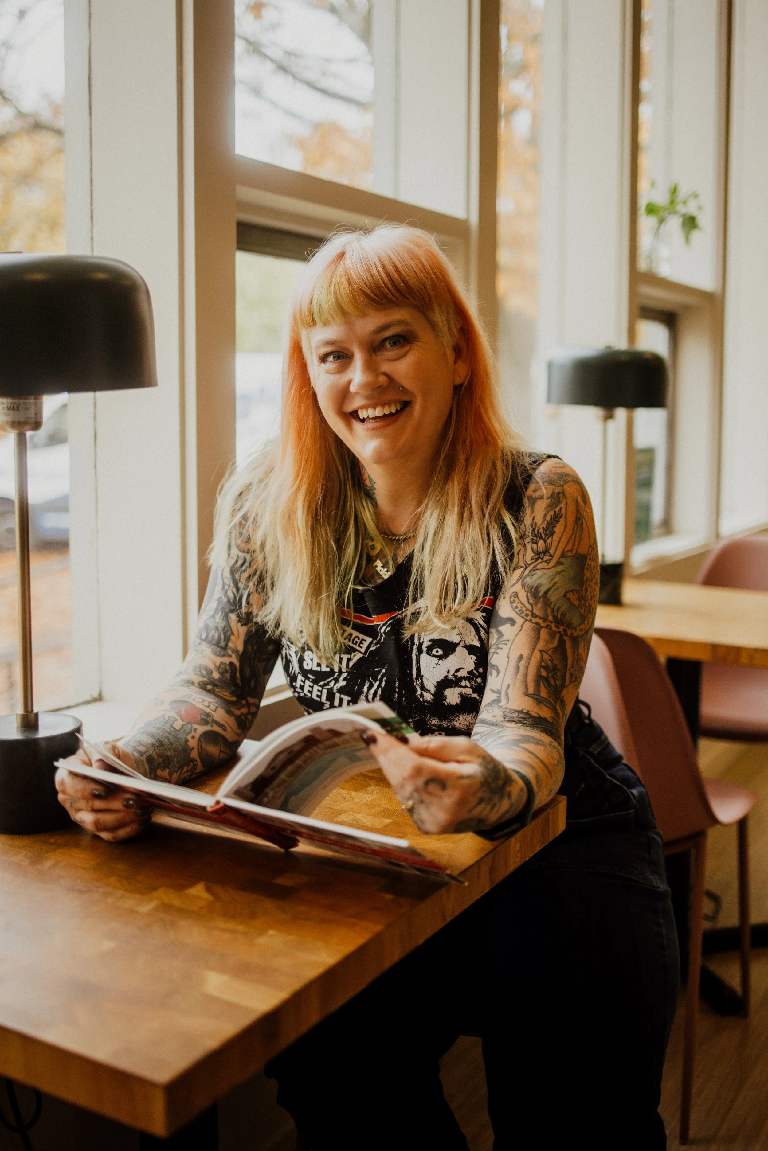 A smiling woman with orange hair and tattoos on her arms sitting at a wooden table in a cafe, holding an open magazine, with large windows behind her showing autumn trees.