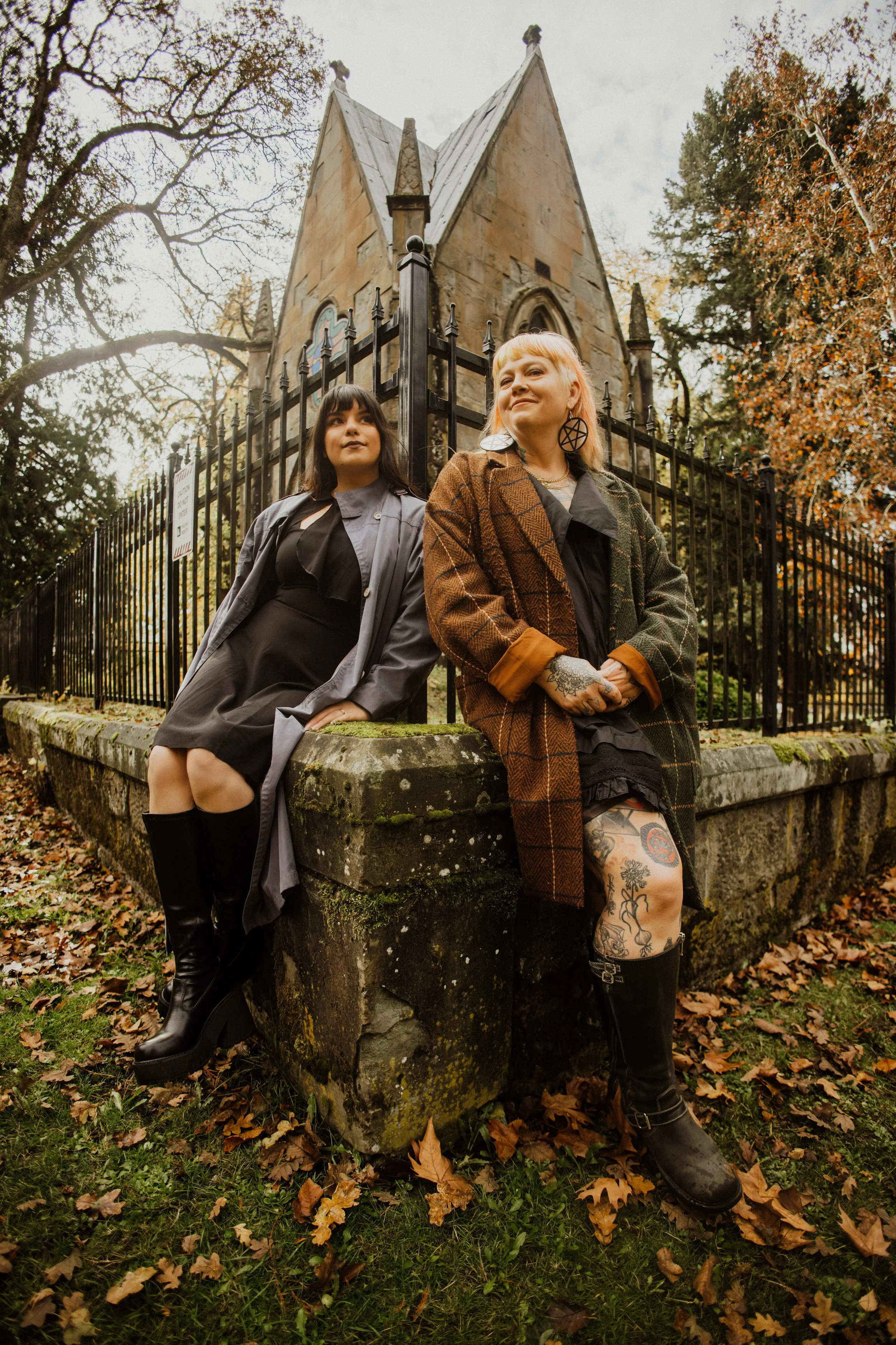 Two women sitting on a moss-covered stone wall outside a church with autumn trees in the background. The woman on the left has dark hair, wearing a black dress and gray trench coat. The woman on the right has blonde hair, tattoos on her legs, and is 