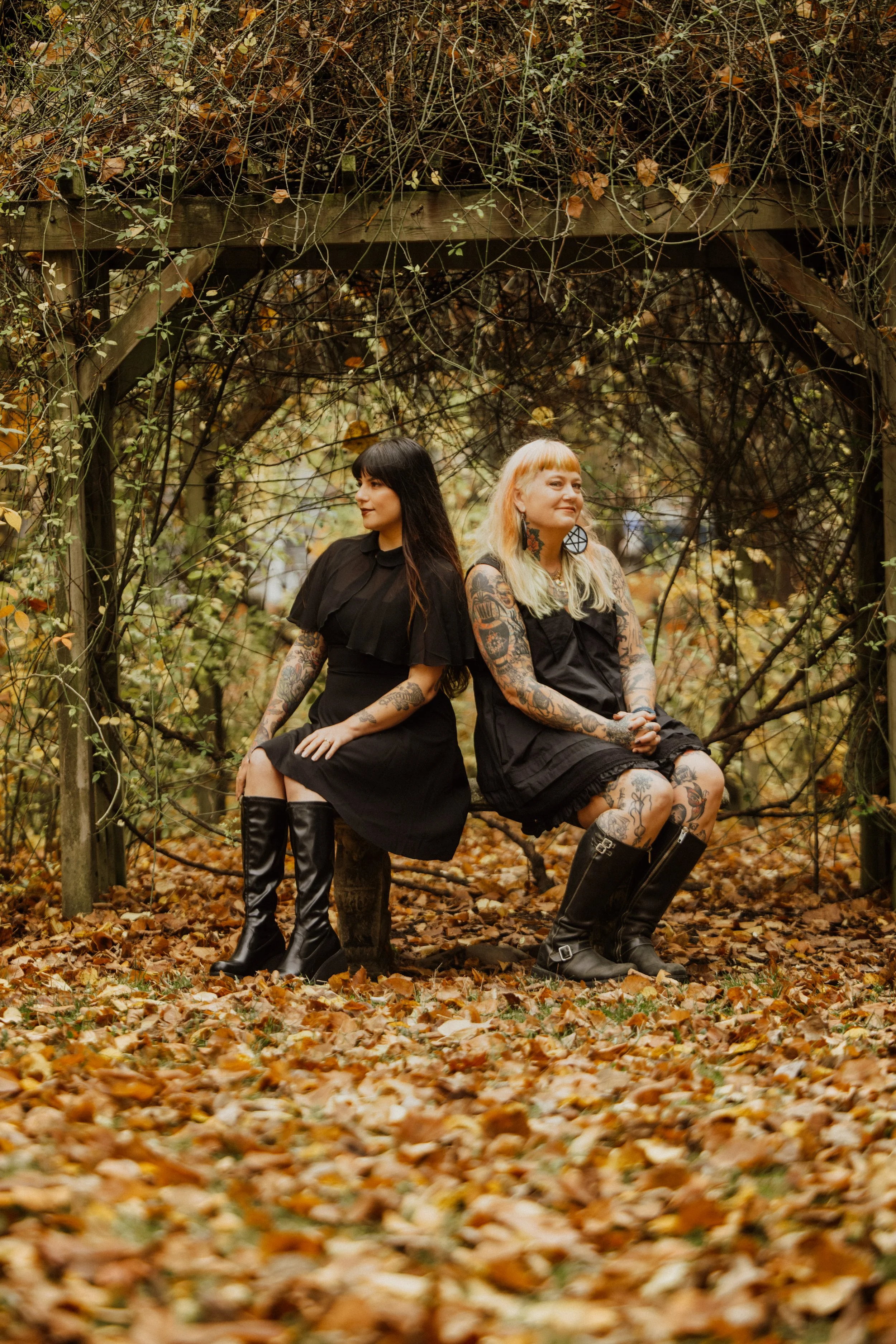 Two women with tattoos sitting on a wooden bench in an autumnal outdoor setting, surrounded by fallen leaves and a rustic wooden arbor covered in vines and foliage.