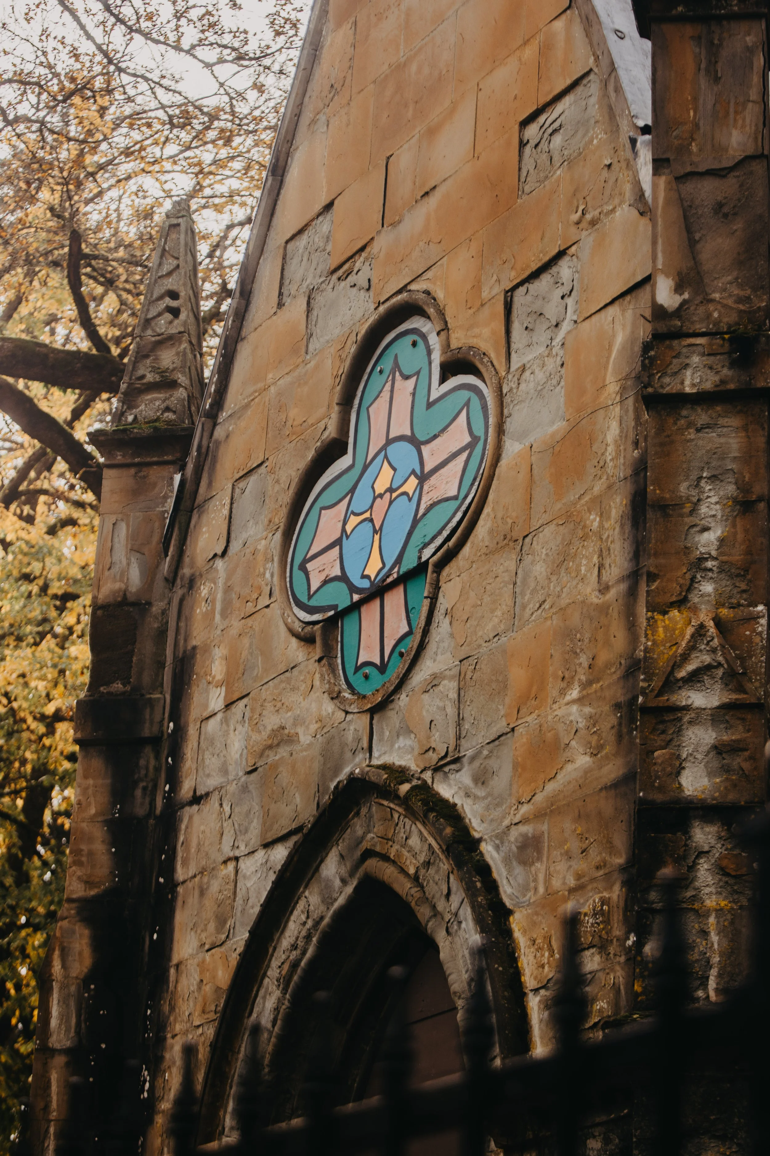 Close-up of a stained glass window on a stone church wall, featuring a floral design with orange, blue, green, and purple colors, and a pointed arch window below it.