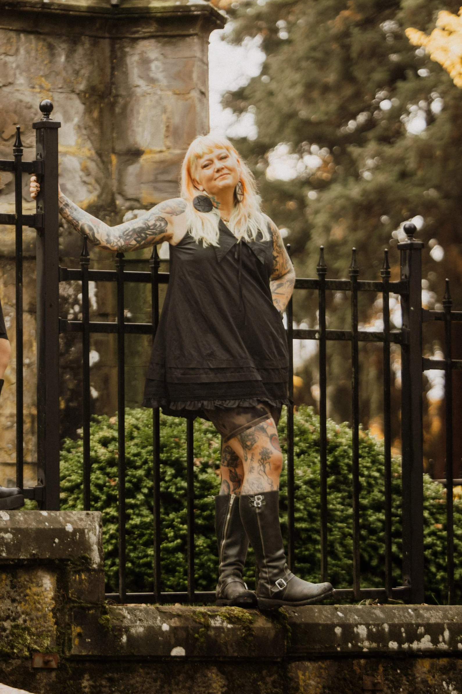A woman with blonde hair and tattoos wearing a black dress and knee-high boots standing outdoors next to a black iron fence