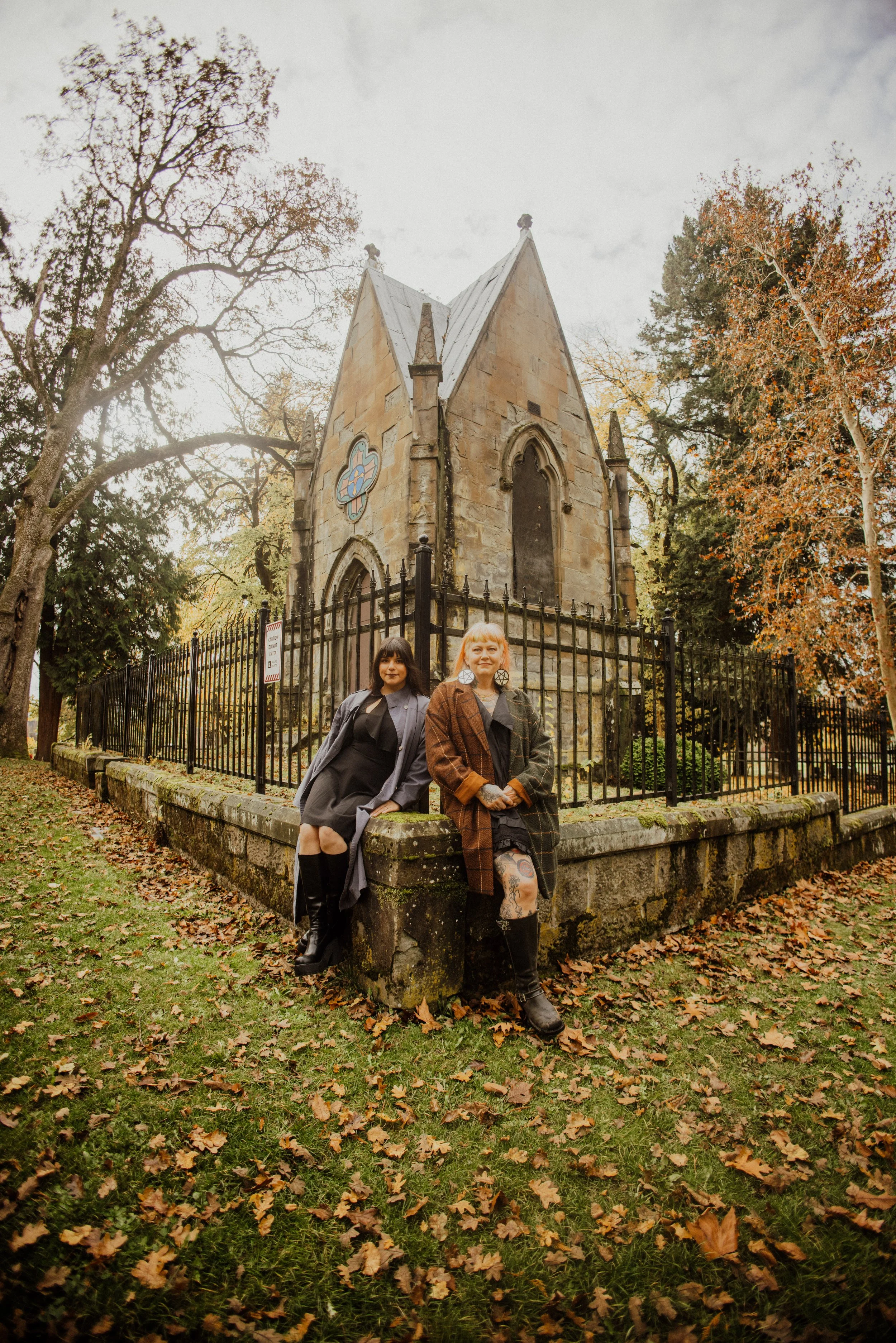 Two women sitting on a stone wall in front of an old stone church with gothic architecture, surrounded by autumn trees with fallen leaves on the ground.