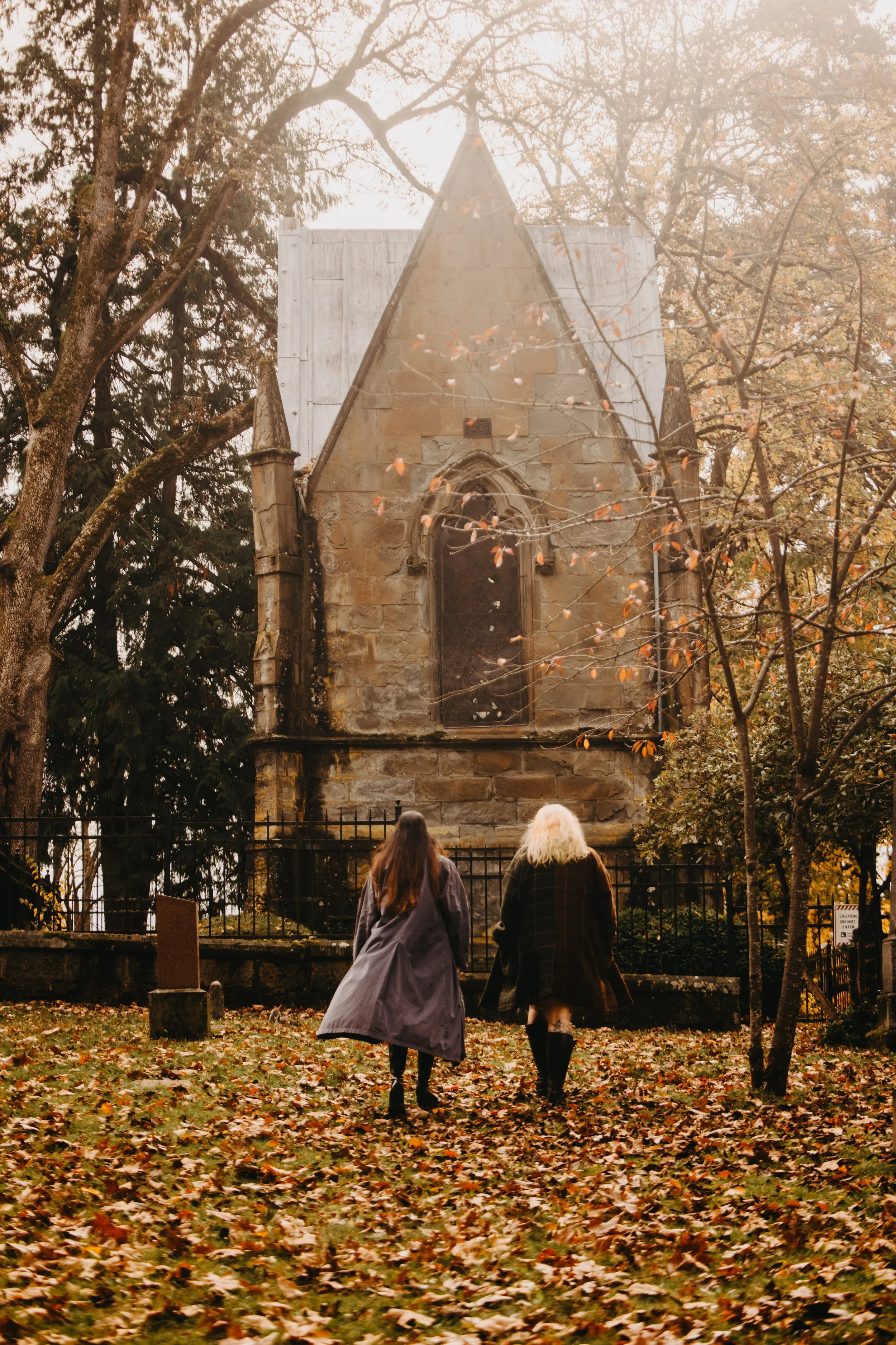 Two women walking towards an old, abandoned church surrounded by fall foliage.