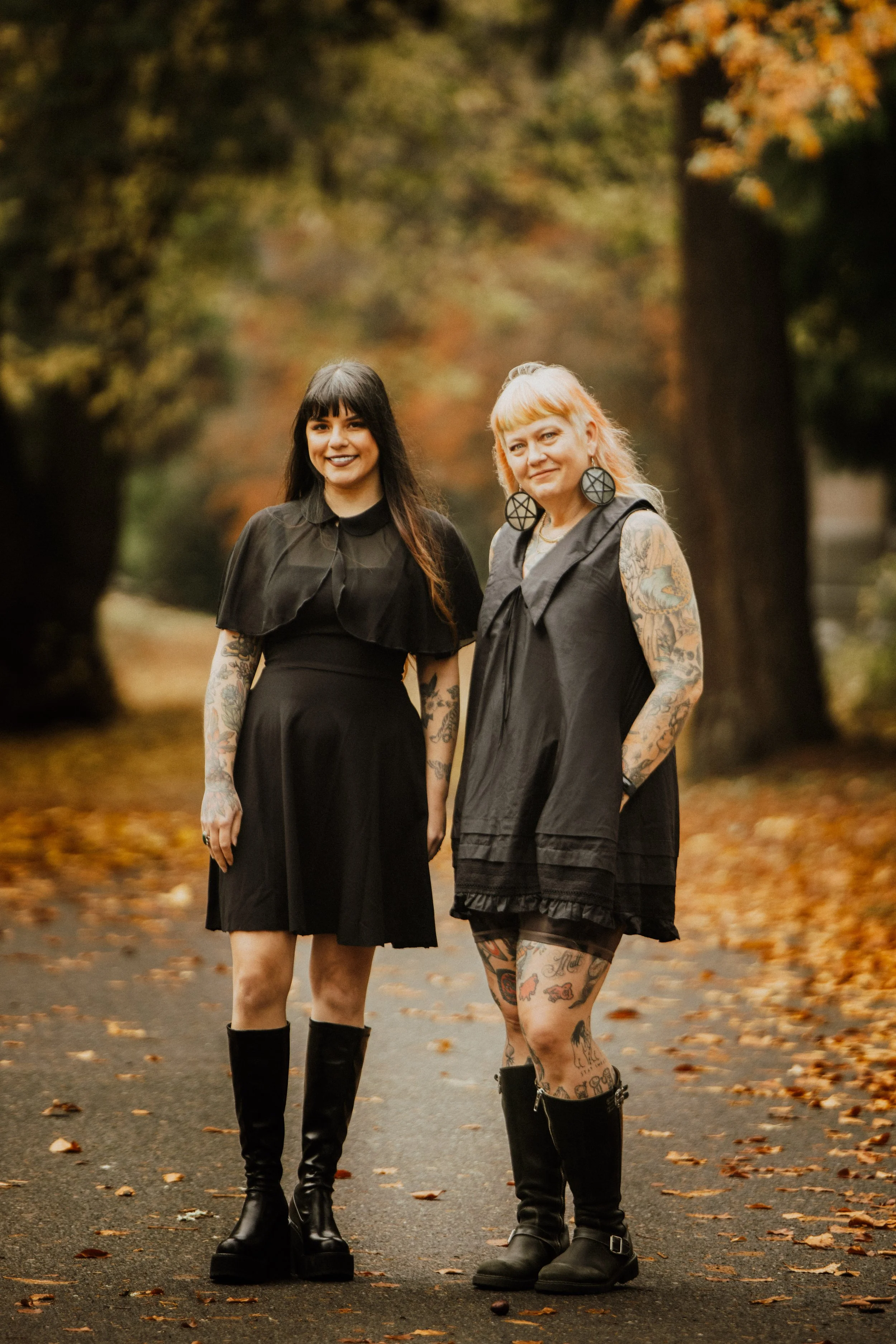 Two women standing on a fall-colored wooded path, both with tattoos and wearing black dresses and knee-high black boots, smiling at the camera.