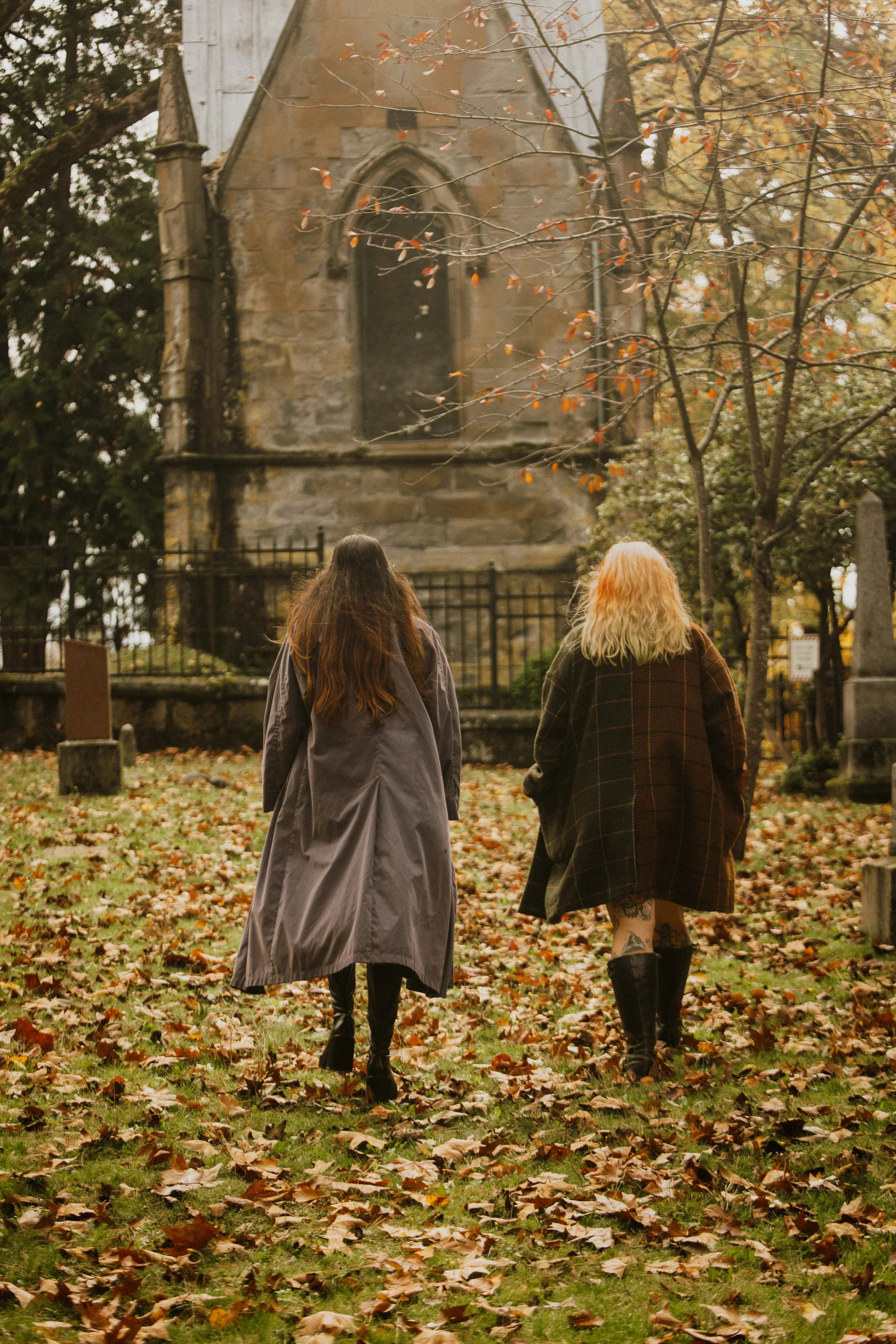 Two women with long hair walking through a park on a fall day, with autumn leaves on the ground, and an old stone church in the background.