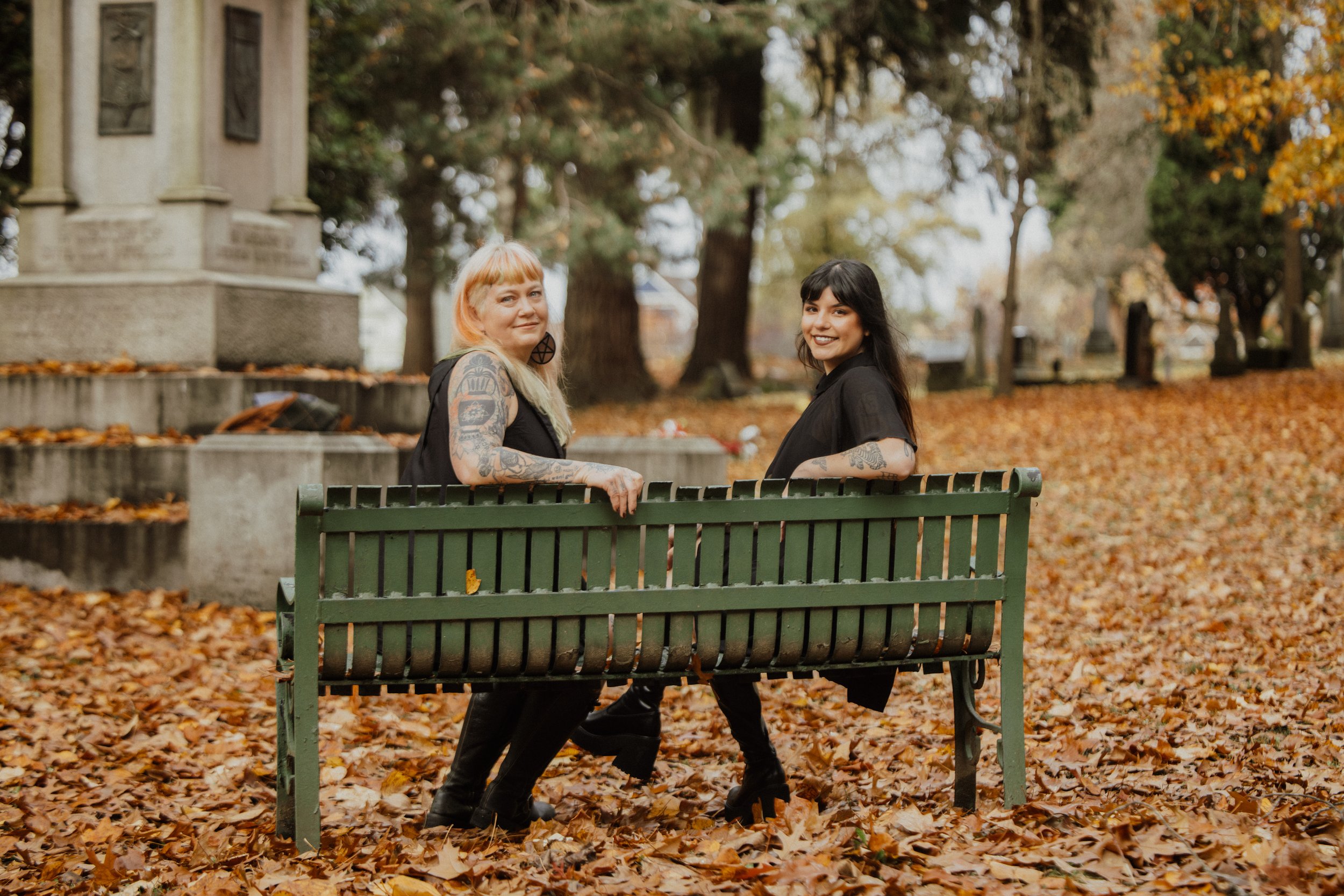 Two women with tattoos sitting on a green park bench in an autumn park with fallen leaves, smiling at the camera.