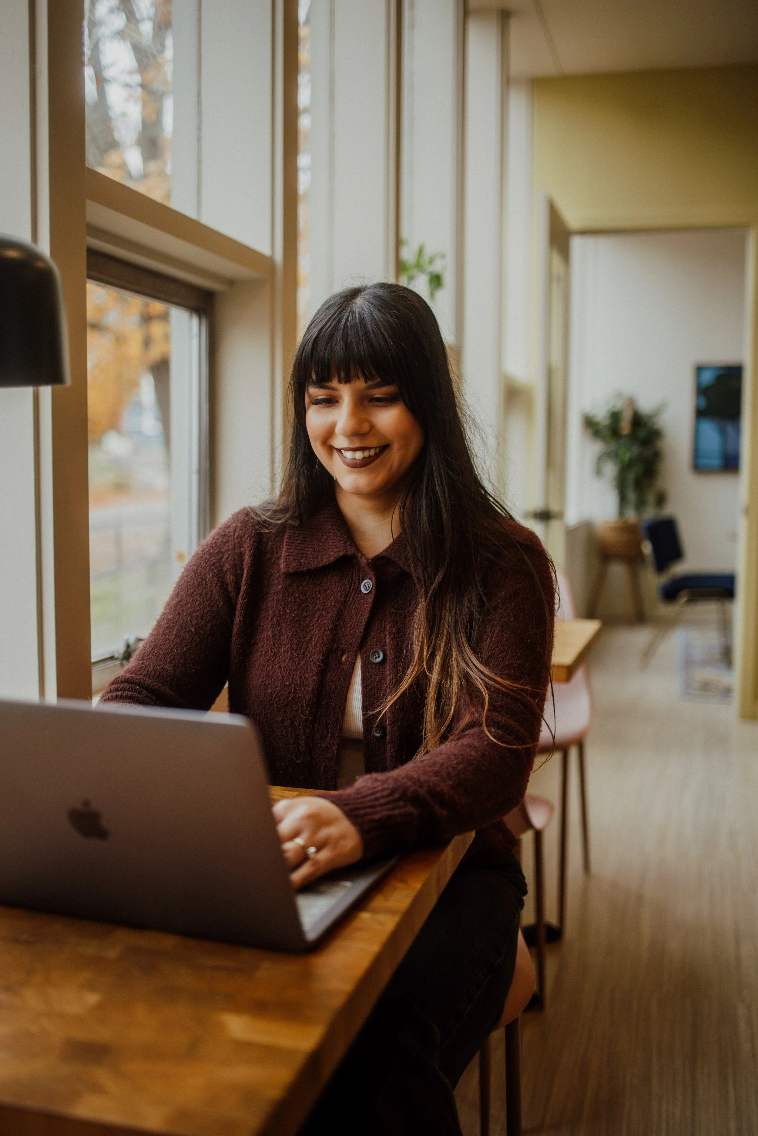 A woman with dark hair and a smile is working on a laptop at a wooden table near a window in a cozy, well-lit room.