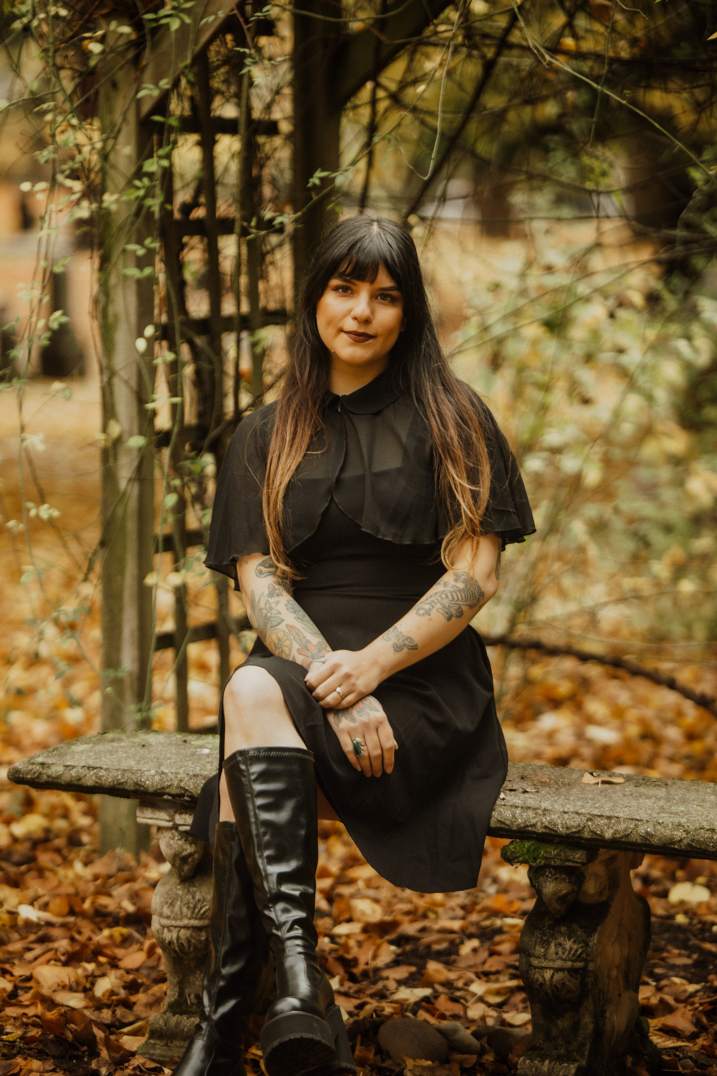 A woman with dark hair and tattoos sitting on a stone bench in an autumnal outdoor setting, wearing a black dress and black leather boots.