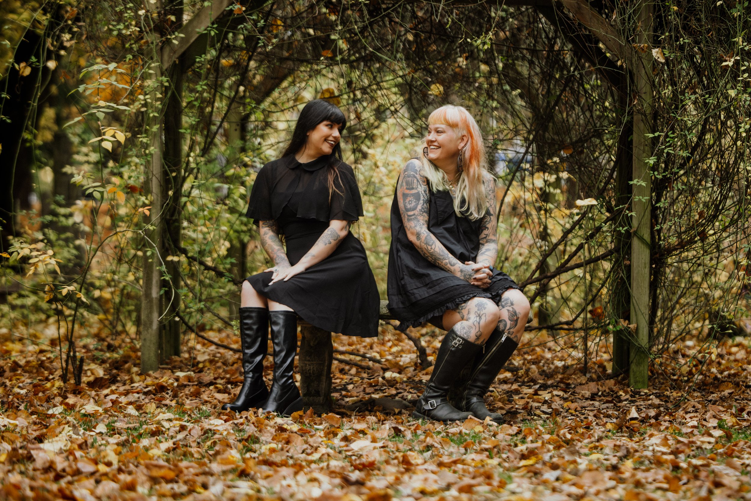 Two women sitting on a bench in an autumn forest, smiling and looking at each other, both wearing black dresses and knee-high boots, with tattoos visible on their arms and legs.