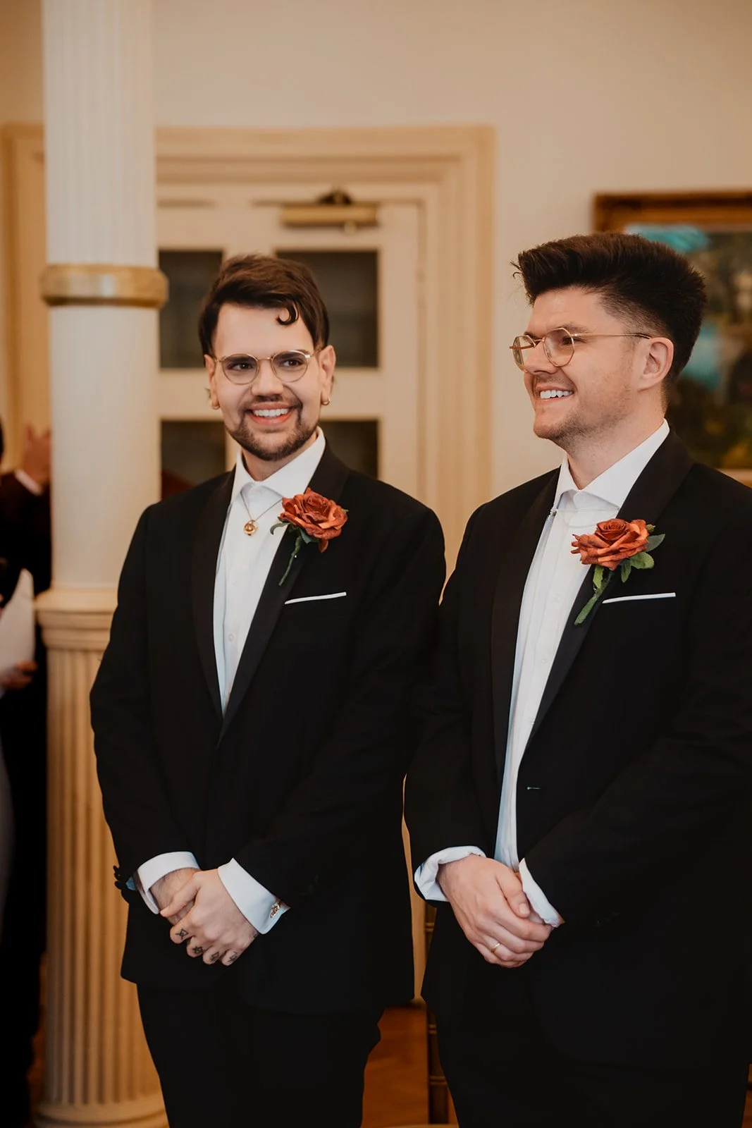 Two men dressed in black tuxedos with white shirts and orange roses on their lapels, standing indoors at a wedding or formal event, smiling and looking happy.