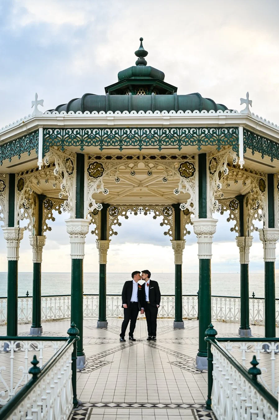 Two men in suits sharing a kiss under a decorative pavilion by the sea.