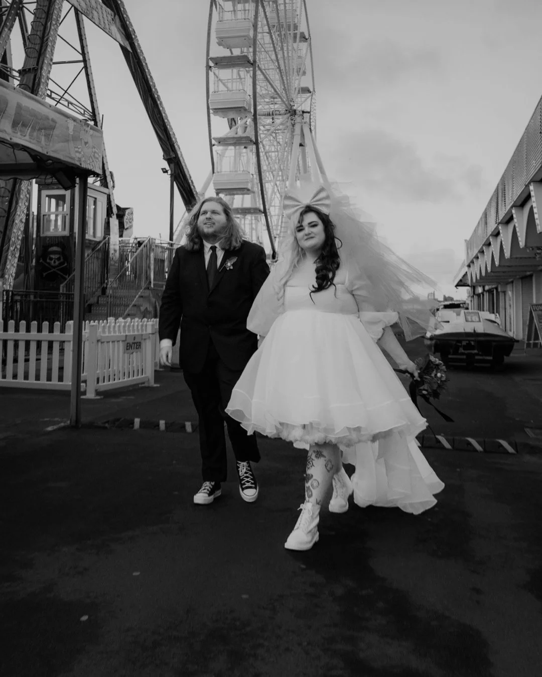 A bride and groom walking at a fairground, with a Ferris wheel and amusement rides in the background, on a cloudy day.