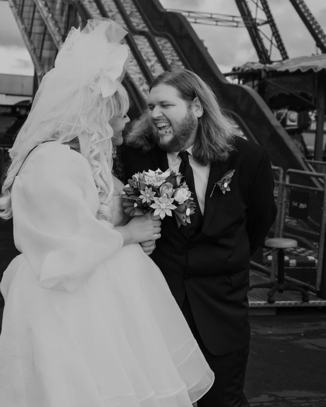 A couple dressed in wedding attire smiling at each other, with the woman holding a bouquet, in front of a carnival ride.