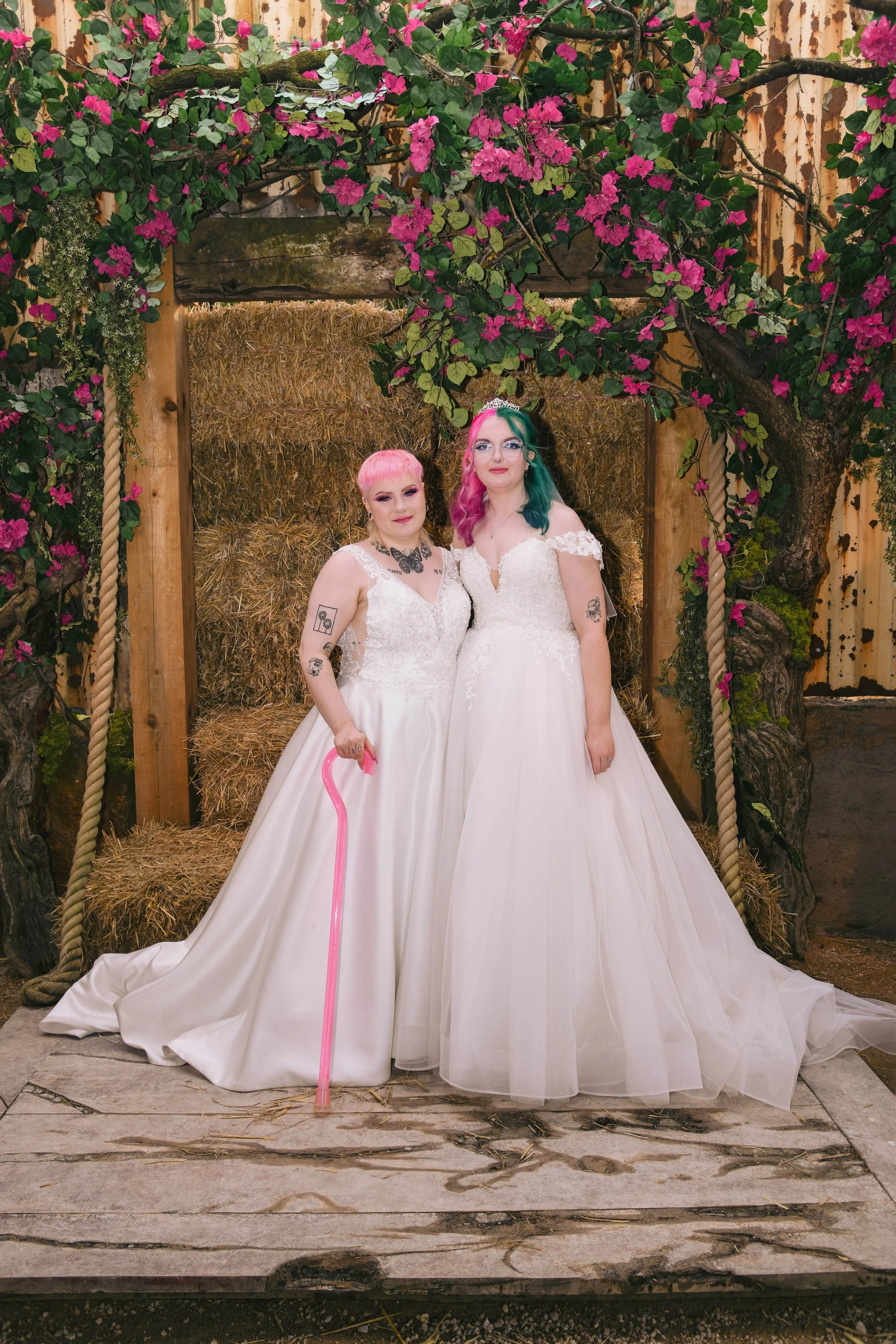 Two women in wedding dresses stand together, surrounded by decorative flowers and a wooden frame, with hay bales in the background.