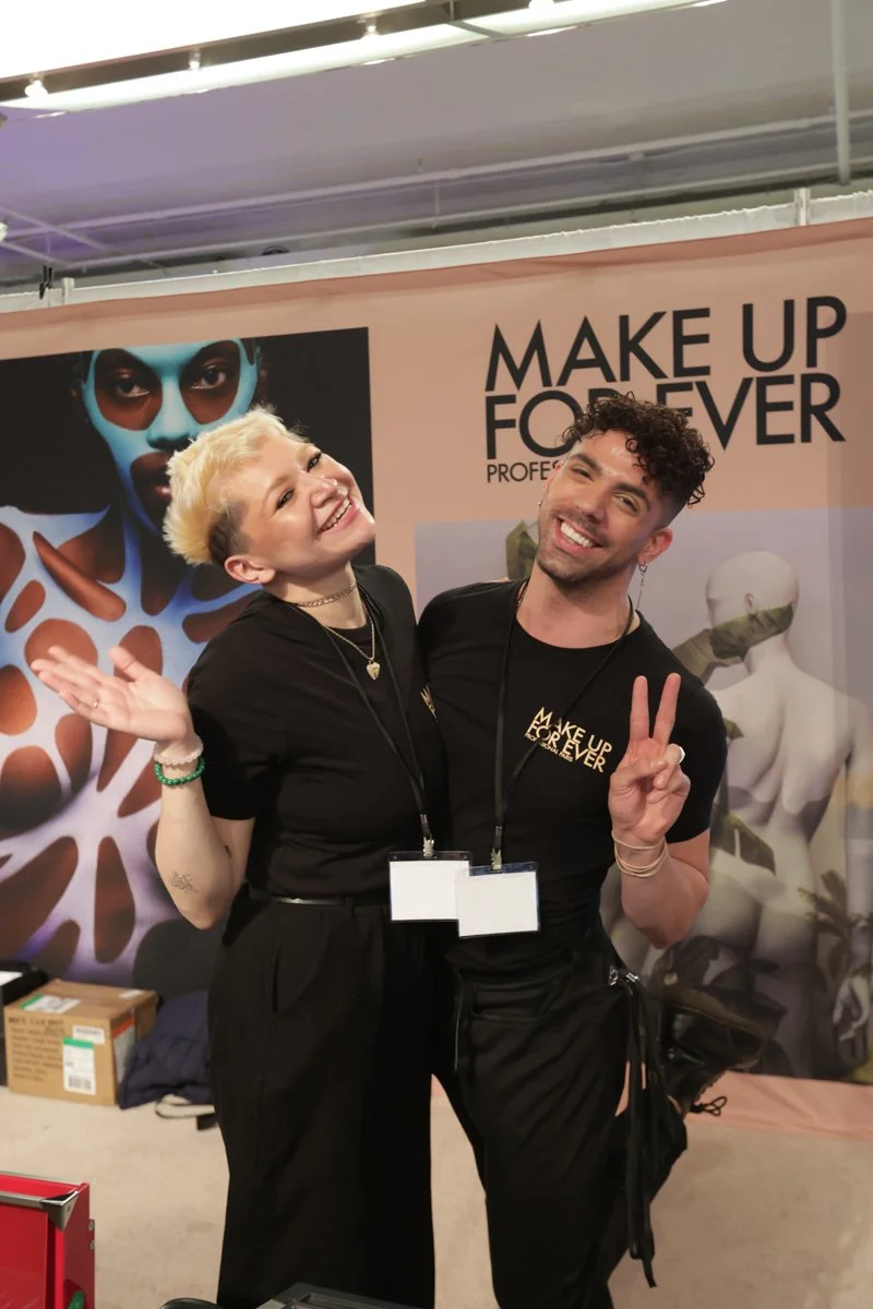 Two beauty professionals pose in front of the Make Up For Ever booth at The Makeup Show.
