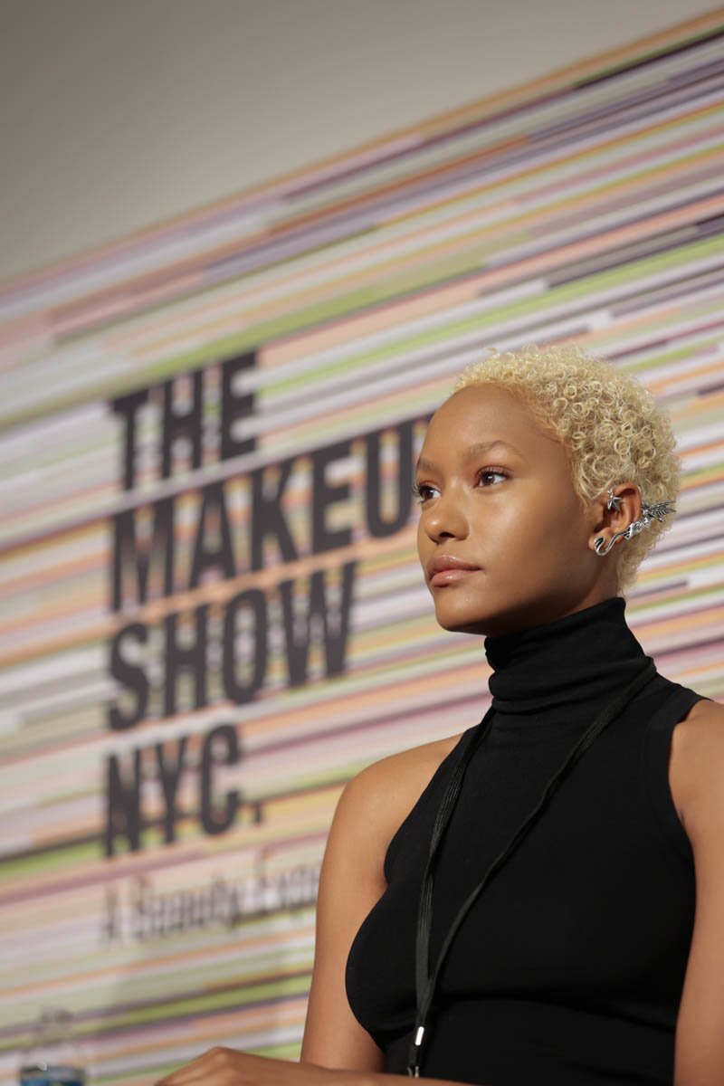 Blonde model in a black outfit posing in front of The Makeup Show NYC event backdrop.