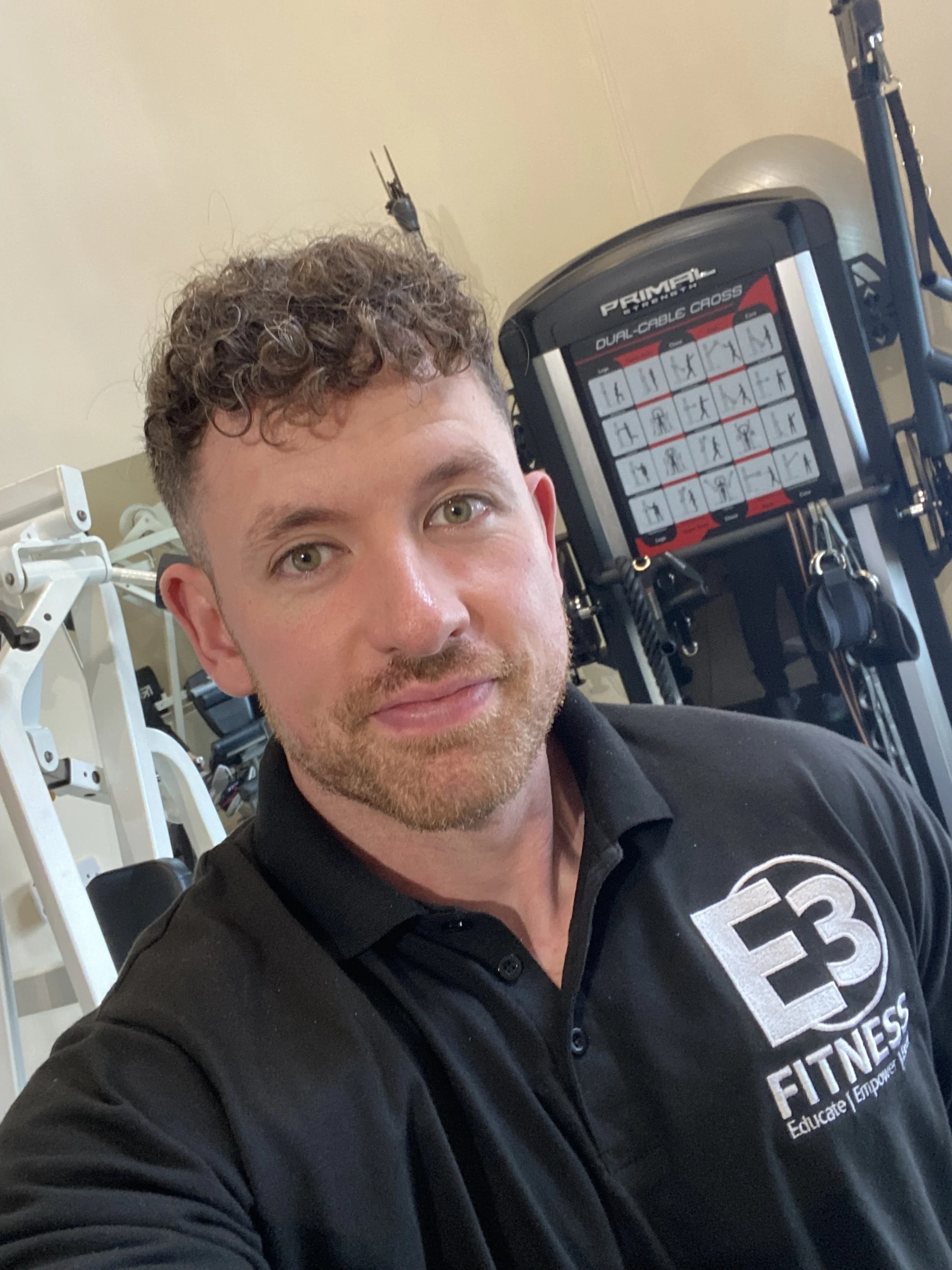 A man with curly brown hair and a beard wearing a black E3 Fitness polo shirt taking a selfie in a gym, with gym equipment in the background.