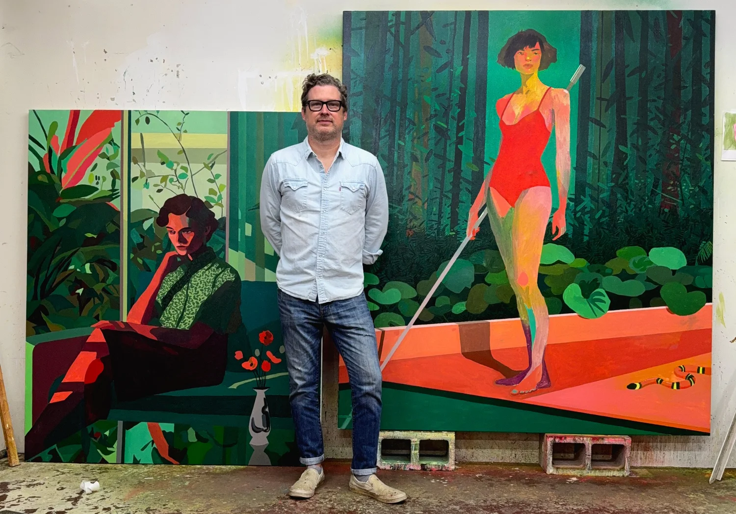 Patrick Puckett standing in his studio in front of two colorful figurative paintings.