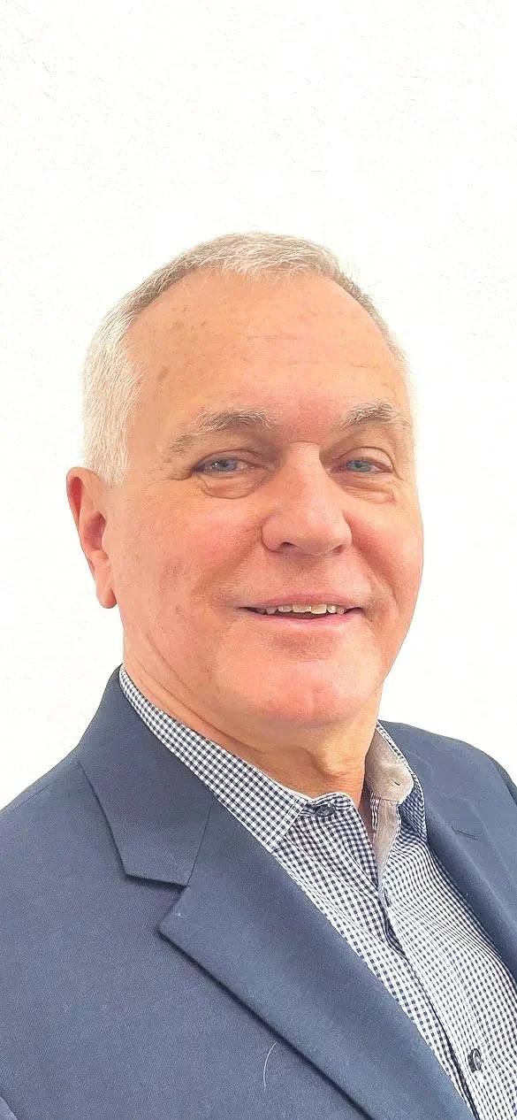 A middle-aged man with short gray hair, wearing a checkered shirt and a dark blazer, smiling with a light-colored plain background.
