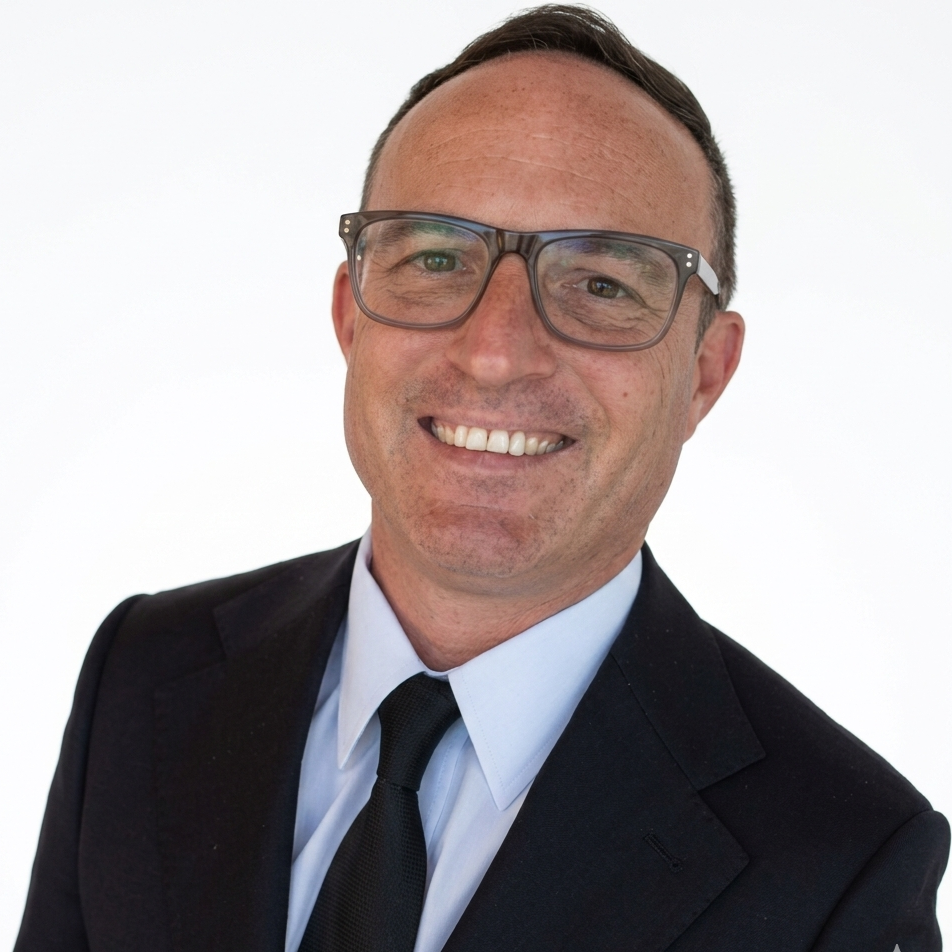 A man wearing glasses, a dark suit, white shirt, and a white tie, smiling and standing against a white background.