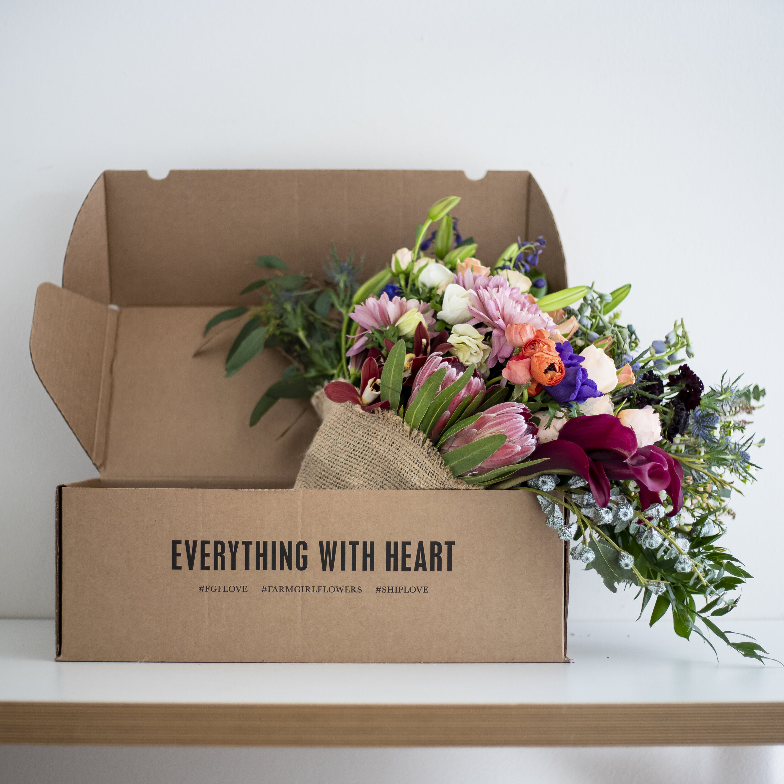 A bouquet of mixed pink, burgundy, and purple flowers wrapped in burlap, sitting in a cardboard box