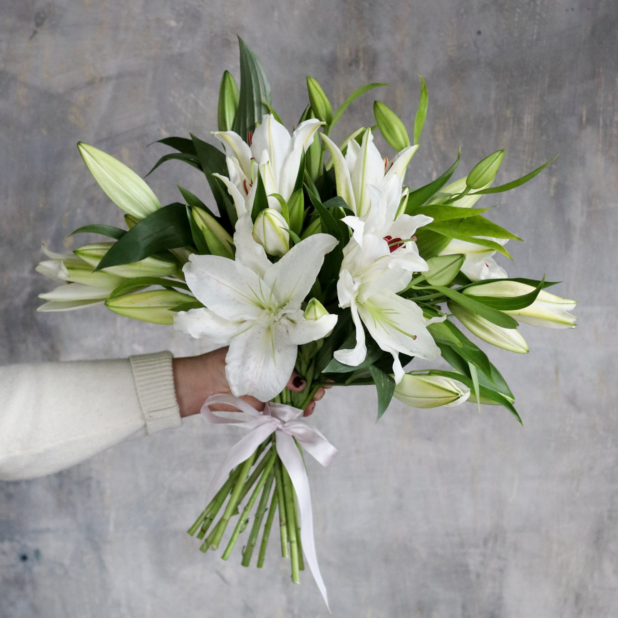 A bouquet of white lilies and greenery tied with a white bow, held up in front of a gray background