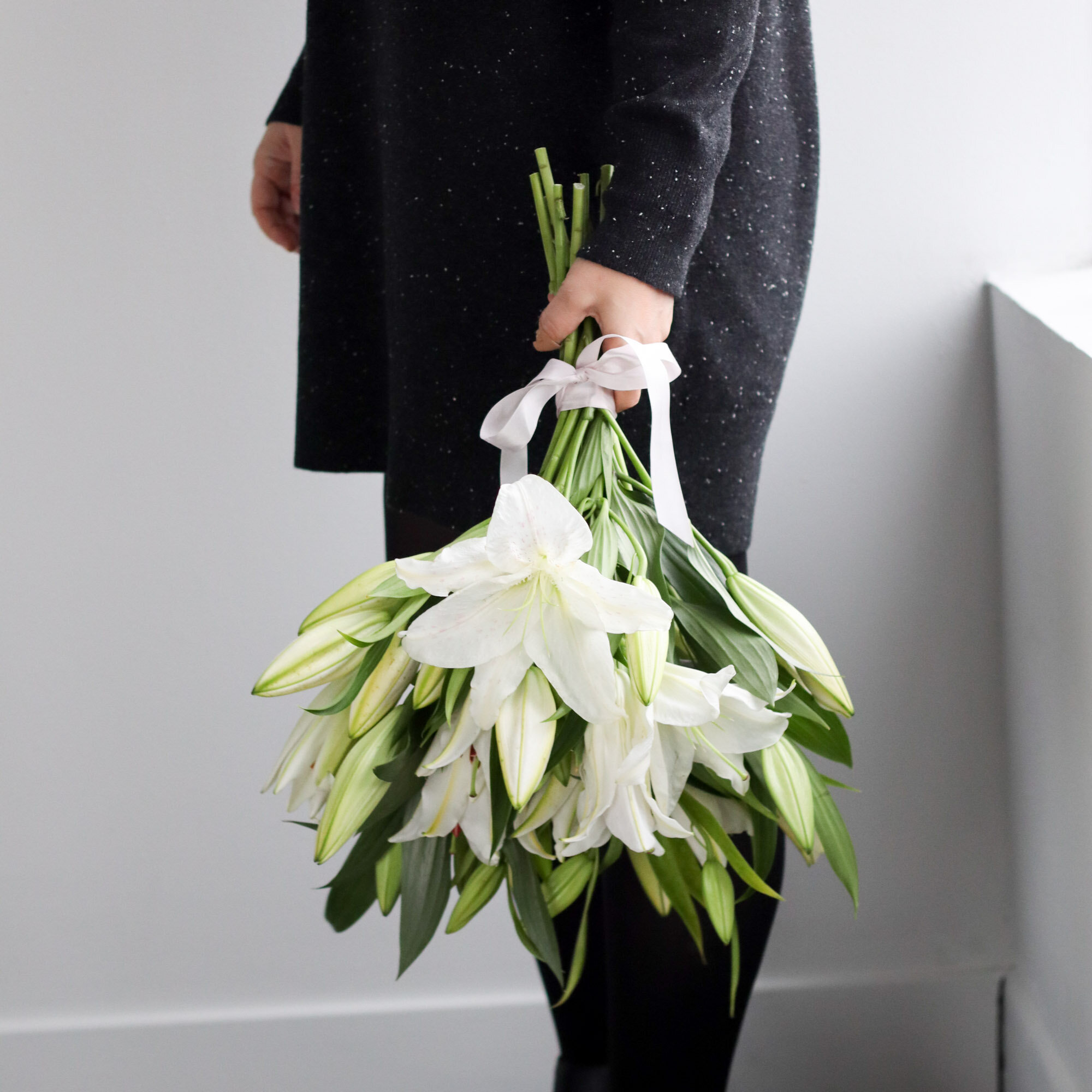 A bouquet of white lilies and greenery tied with a white bow, held upside down beside a person