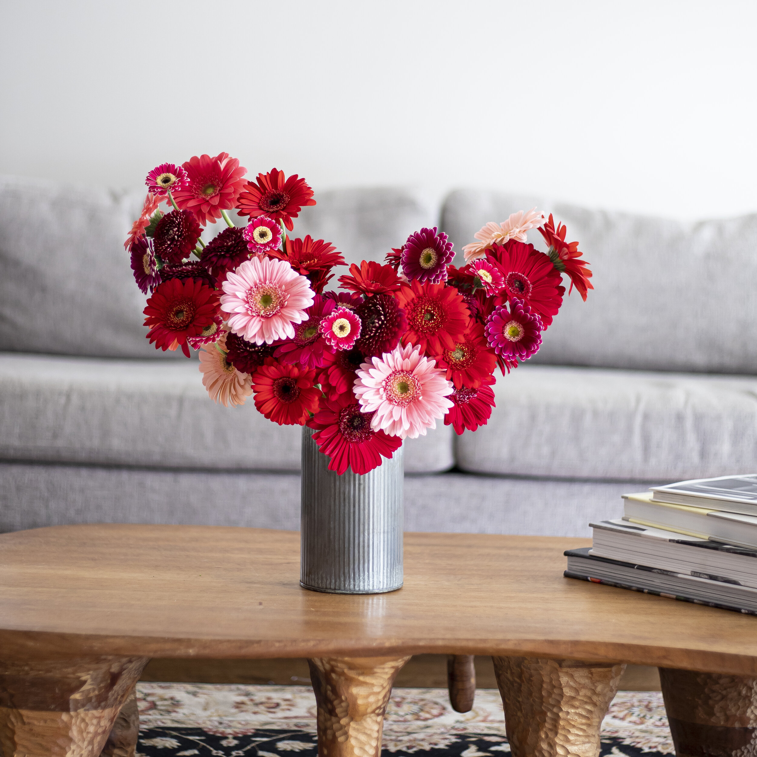 A bouquet of red and pink daisies in a tin vase on a wooden table near a couch