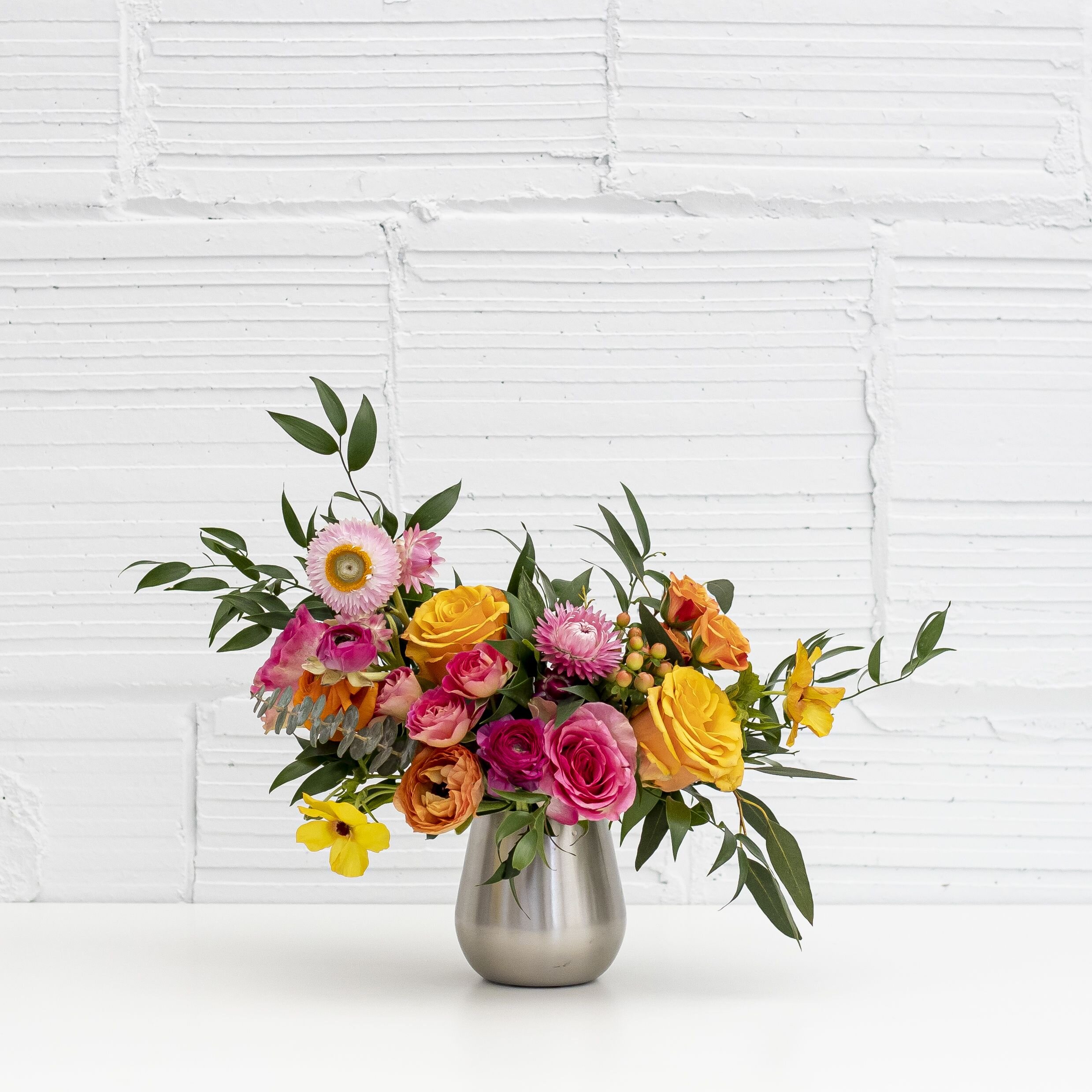 A floral arrangement sitting on a table top with bright pink flowers, yellow flowers, and greenery in a silver vase.