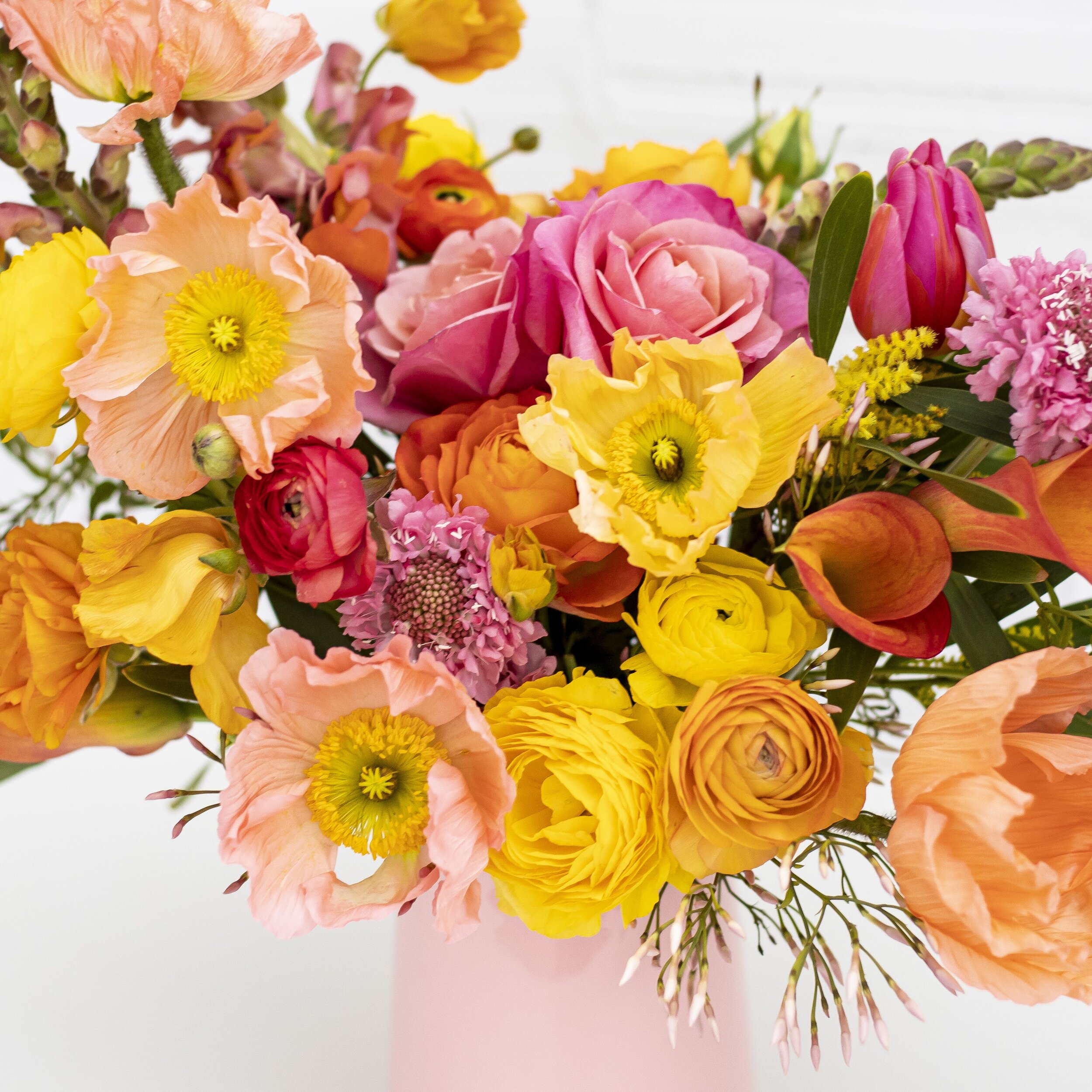 A close up of yellow flowers, pink flowers, and peach colored flowers sitting on a table top in a light pink vase.