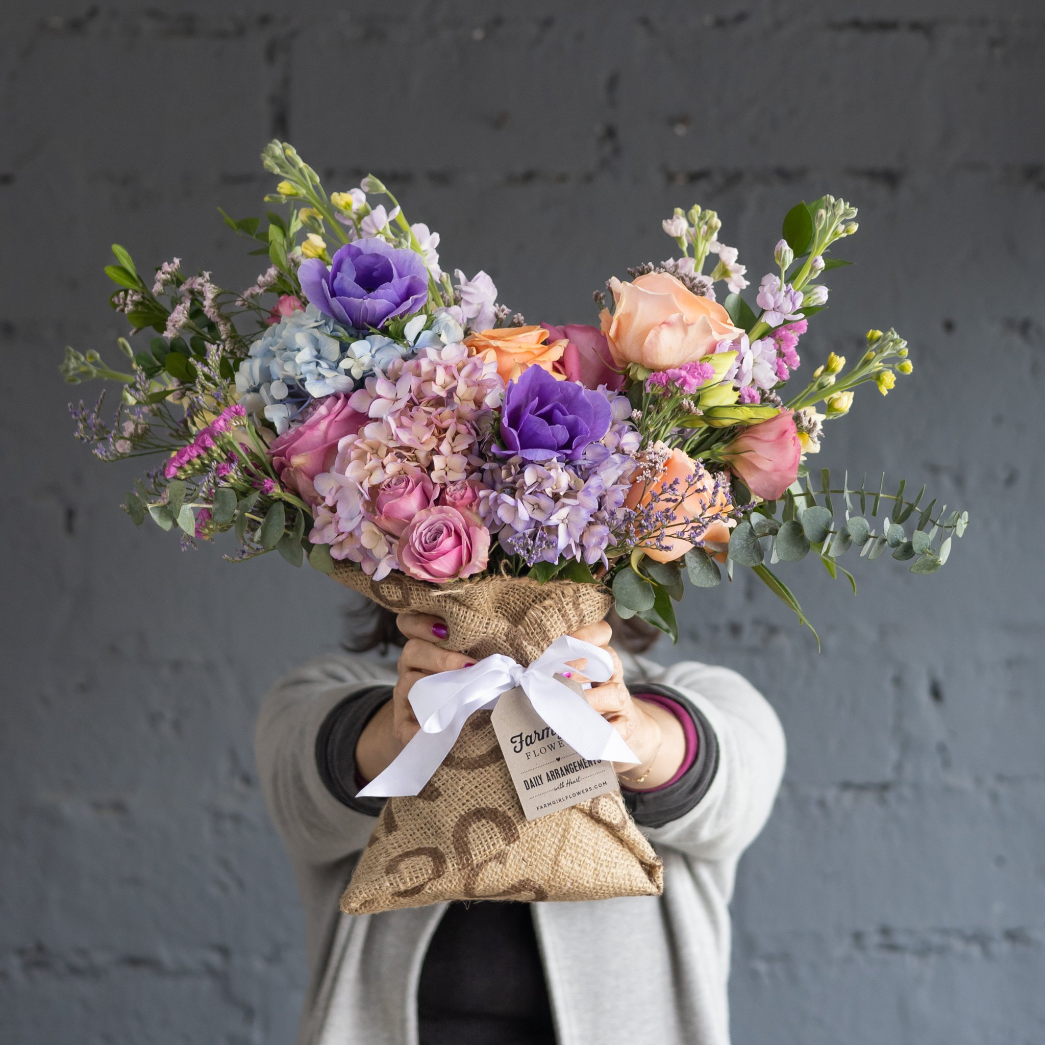A pink and purple floral bouquet wrapped in burlap being held up with two hands, covering the person's face, in front of a grey brick wall.
